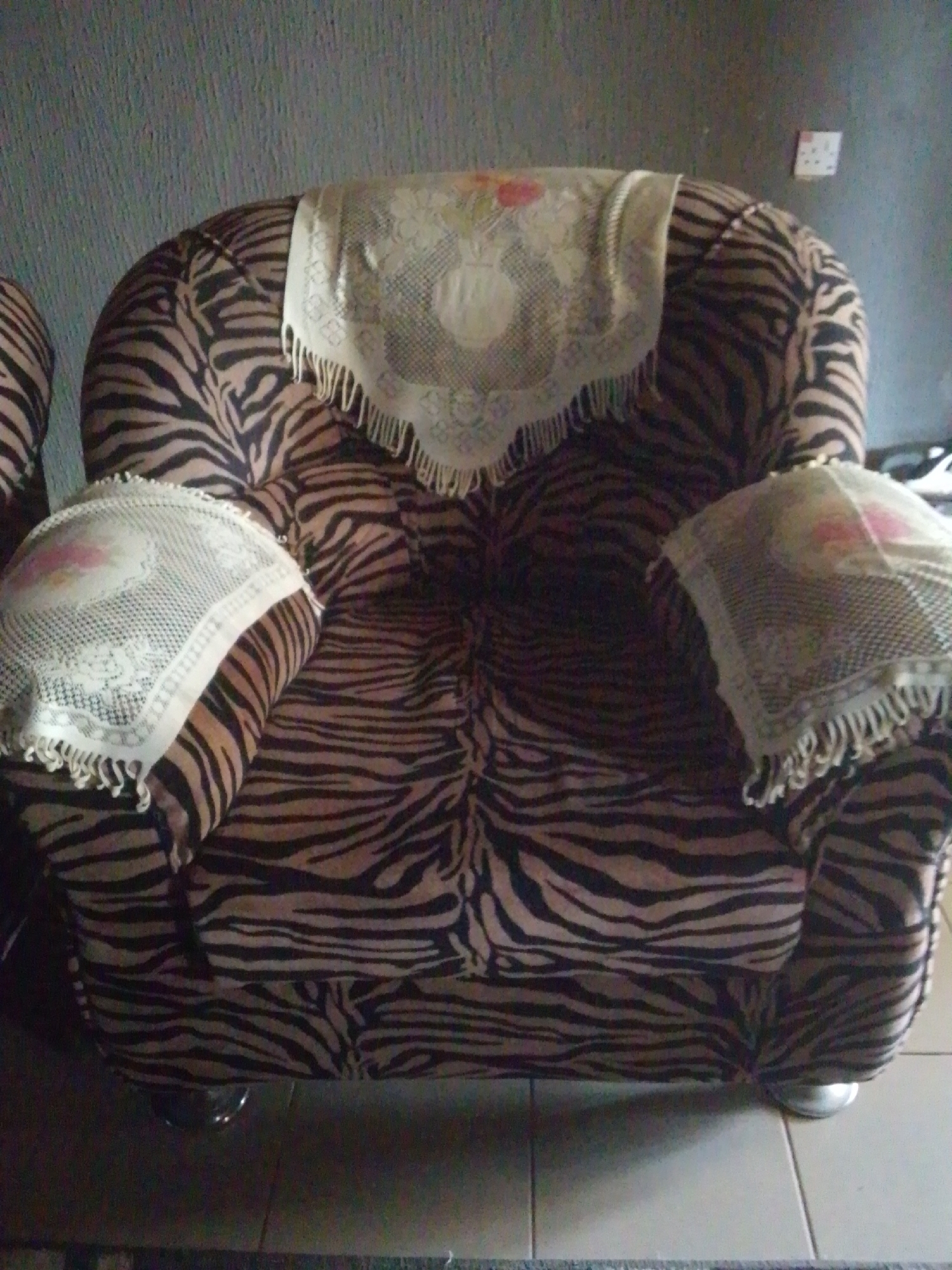 The image shows a close-up, eye-level view of a sofa with a black and brown zebra print upholstery. The sofa appears to be made of fabric and has a plush, rounded design. It is adorned with two decorative white lace doilies, one placed centrally on the back of the sofa and another on the left armrest. These doilies have a floral pattern and fringed edges, adding a touch of traditional decor. The sofa has silver-colored feet, barely visible at the bottom. The background is a textured, gray wall, and on the right side of the wall, a white electrical outlet with two sockets is visible. The lighting suggests an indoor setting, likely during the day, with no specific time cues. The scene is static, with no activity taking place, simply a portrayal of the sofa as an object within a room. The overall impression is one of a well-furnished, domestic interior. Given the provided location context of Jalingo, Nigeria, this domestic scene is situated in that region.