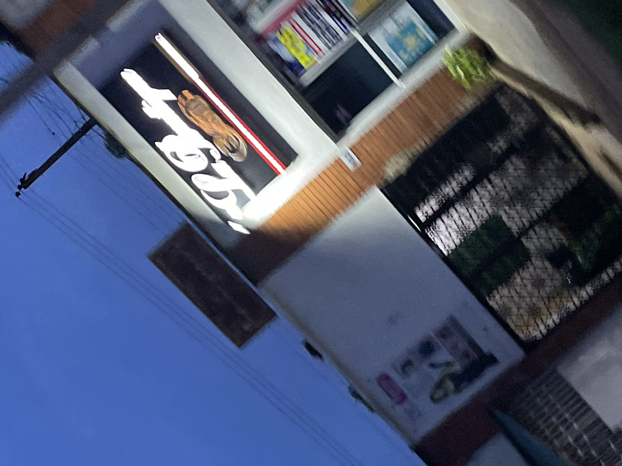 The image depicts a low-angle, motion-blurred view of a commercial building at dusk, with the sky a deep blue. The building has a brick facade and a white upper section where a prominent sign with neon lettering is visible. The sign appears to spell out "KG Bar" with a stylized image of a horseshoe. Below the sign, framed advertisements or posters are displayed in a window, one of which features a woman in dark clothing. A chain-link fence with a black gate is in the foreground, and a potted plant with green foliage is visible to the right. The overall impression is that of a street-level establishment in an urban environment. The image is not specific enough to determine the exact location in Jos, Nigeria, beyond it being a commercial area.