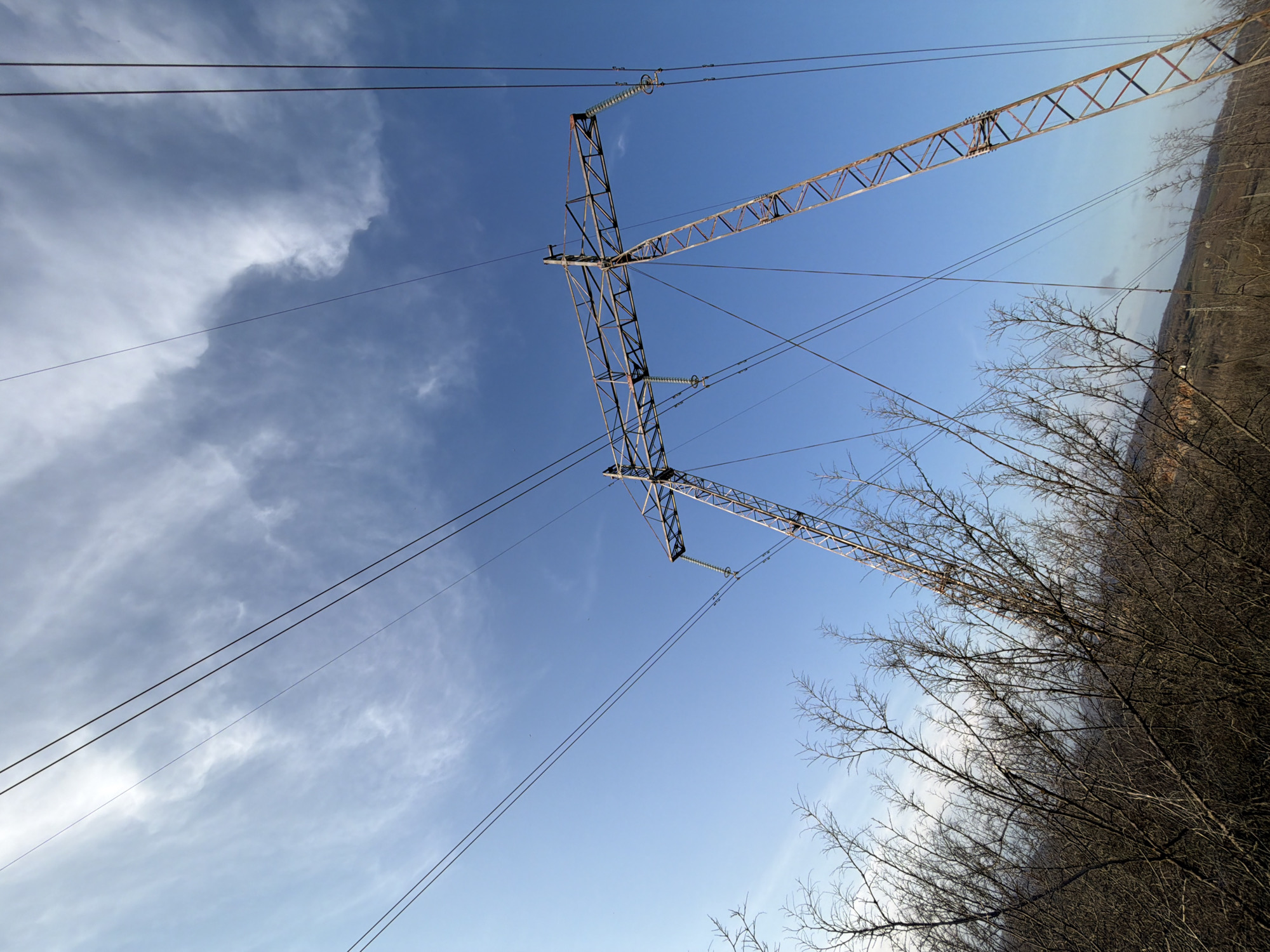 The image shows a low-angle view of a metal lattice electricity pylon against a backdrop of a light blue sky with scattered clouds. Several high-voltage power lines stretch across the frame. To the right, bare branches of trees and bushes are visible, suggesting a rural or semi-rural environment. Based on the prompt, the location is Karlukovo, Bulgaria, implying this is part of the local electrical infrastructure. There are no people visible in the image.