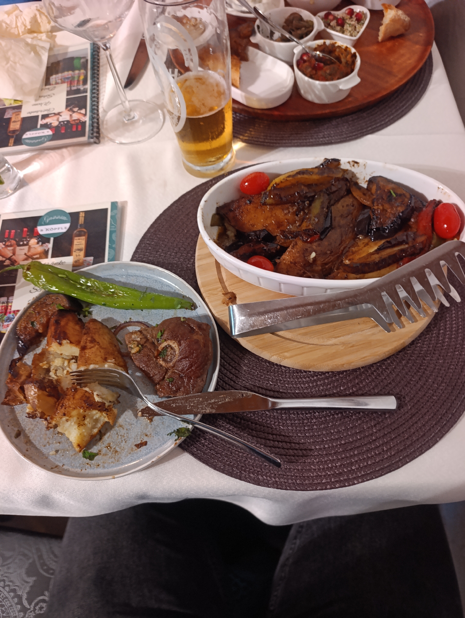 The image shows a close-up view of a table at a restaurant, likely in Reutlingen, Germany, given the presence of a menu for "Gasthaus S'Köpfle". The table is set with a white tablecloth, a brown placemat, and cutlery. On the placemat are two plates of food. The plate in the foreground contains roasted potato wedges, a piece of lamb, and a green pepper. A fork and knife are also present. Behind this plate, on the placemat, is a white oval dish filled with a cooked dish of eggplant and tomatoes, with two small cherry tomatoes visible on the rim. A pair of silver tongs rests on a wooden board next to the oval dish. To the left of the food, there are two menus. A glass of beer is visible in the upper right portion of the image, and another empty wine glass is to its left. In the background, a round wooden tray holds several small white bowls containing appetizers, and a piece of bread. The overall scene suggests a meal in progress at a restaurant.