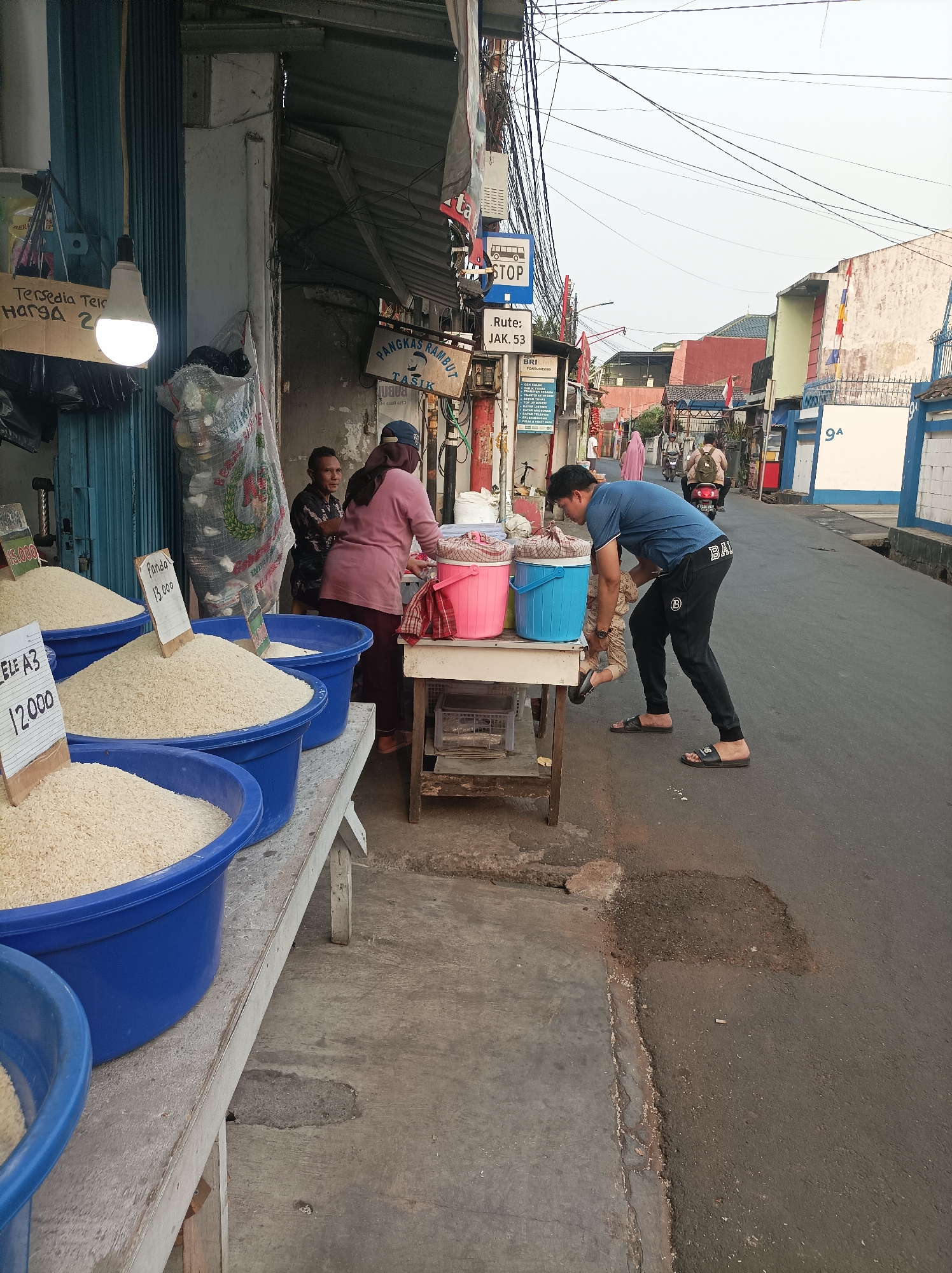 An outdoor street-level view captures a bustling market scene in West Jakarta, Indonesia. In the foreground, several large blue tubs are filled with mounds of white rice, each with a price tag indicating the cost per unit. A wooden table displays two large buckets, one pink and one blue, filled with a dark liquid, possibly water or another beverage. A man in a blue t-shirt and black pants is bent over, interacting with a child wearing a beige and brown patterned outfit. Two women are visible in the background, one in a pink hijab and sweater, and another wearing a dark patterned top. The market stalls are set up along a paved street, with buildings and overhead wires lining the background. The atmosphere suggests a typical day in a vibrant local marketplace.