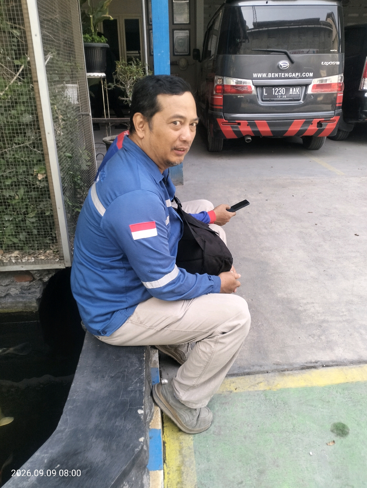 A middle-aged Indonesian man with short, dark hair and a mustache is sitting on a dark gray concrete ledge outdoors, facing slightly left and looking at the camera. He is wearing a blue long-sleeved work shirt with reflective stripes on the sleeves and shoulders, a small Indonesian flag patch on his left sleeve, and light-colored trousers. He has a black bag slung over his right shoulder and is holding a black smartphone in his right hand. To the left of the man, there is a metal-framed enclosure with a mesh wire covering, containing green plants. Behind the man, a dark gray van is parked, with its rear visible. The van has a license plate that reads "L 1230 AAK" and displays a website address "WWW.BENTENGAPI.COM". The ground is paved with a light gray concrete surface, with yellow and green painted lines. The scene appears to be in an outdoor area, possibly a parking lot or a service area, in Bambe, Indonesia, based on the license plate format and the presence of tropical vegetation. The overall lighting suggests it is daytime, possibly late afternoon.