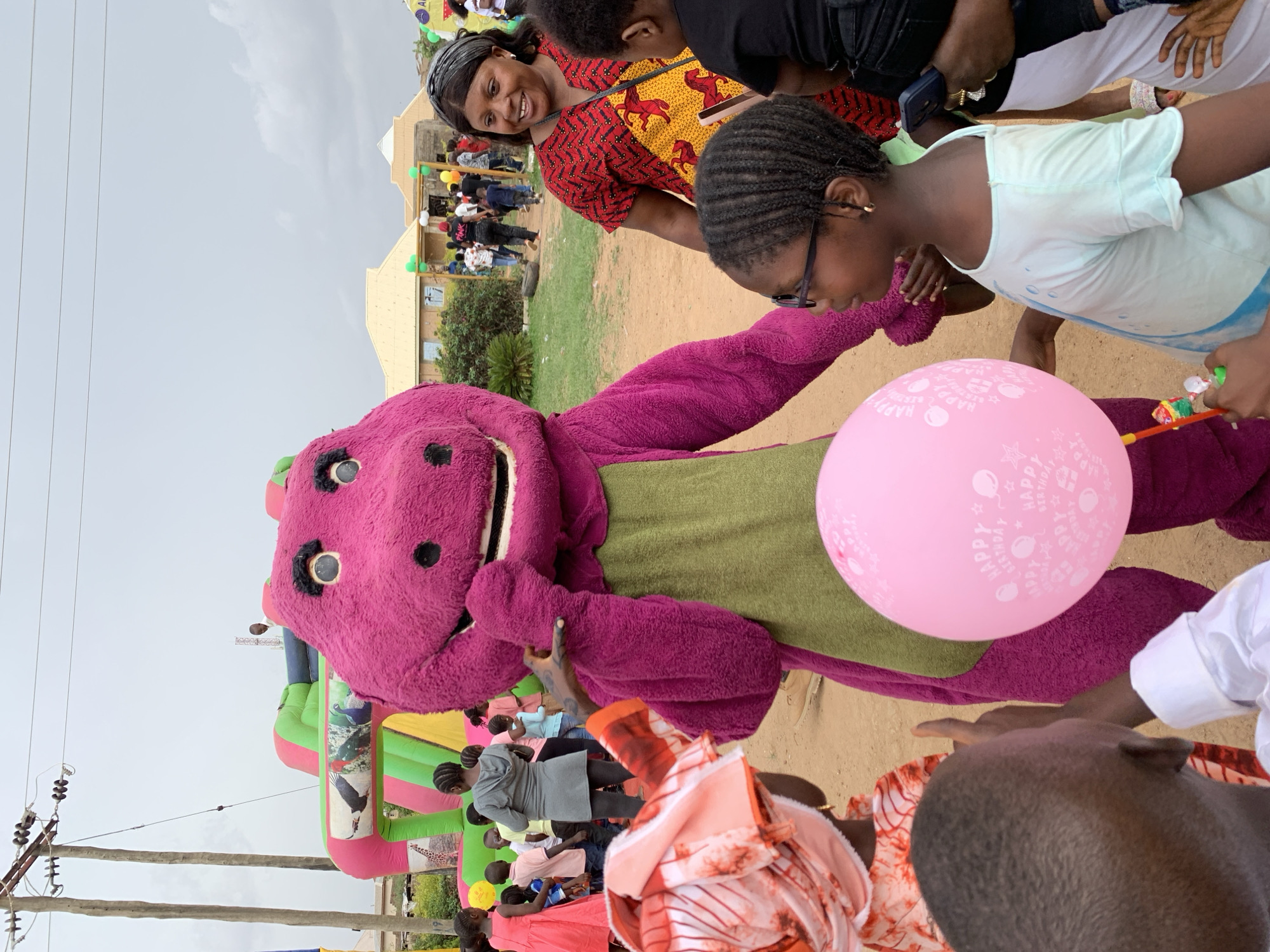 A high-angle medium shot shows a gathering of people interacting with a Barney the Dinosaur mascot, potentially at an event in Jos, Nigeria. Children and adults surround the mascot. One child in a light blue shirt touches a pink "Happy Birthday" balloon held by the Barney mascot. An adult woman with dark hair and a colorful dress looks on with a smile. In the background, additional people are gathered near a structure, possibly a building. A colorful inflatable structure suggests a festive environment. Power lines are visible against a cloudy sky. The ground appears to be sandy.