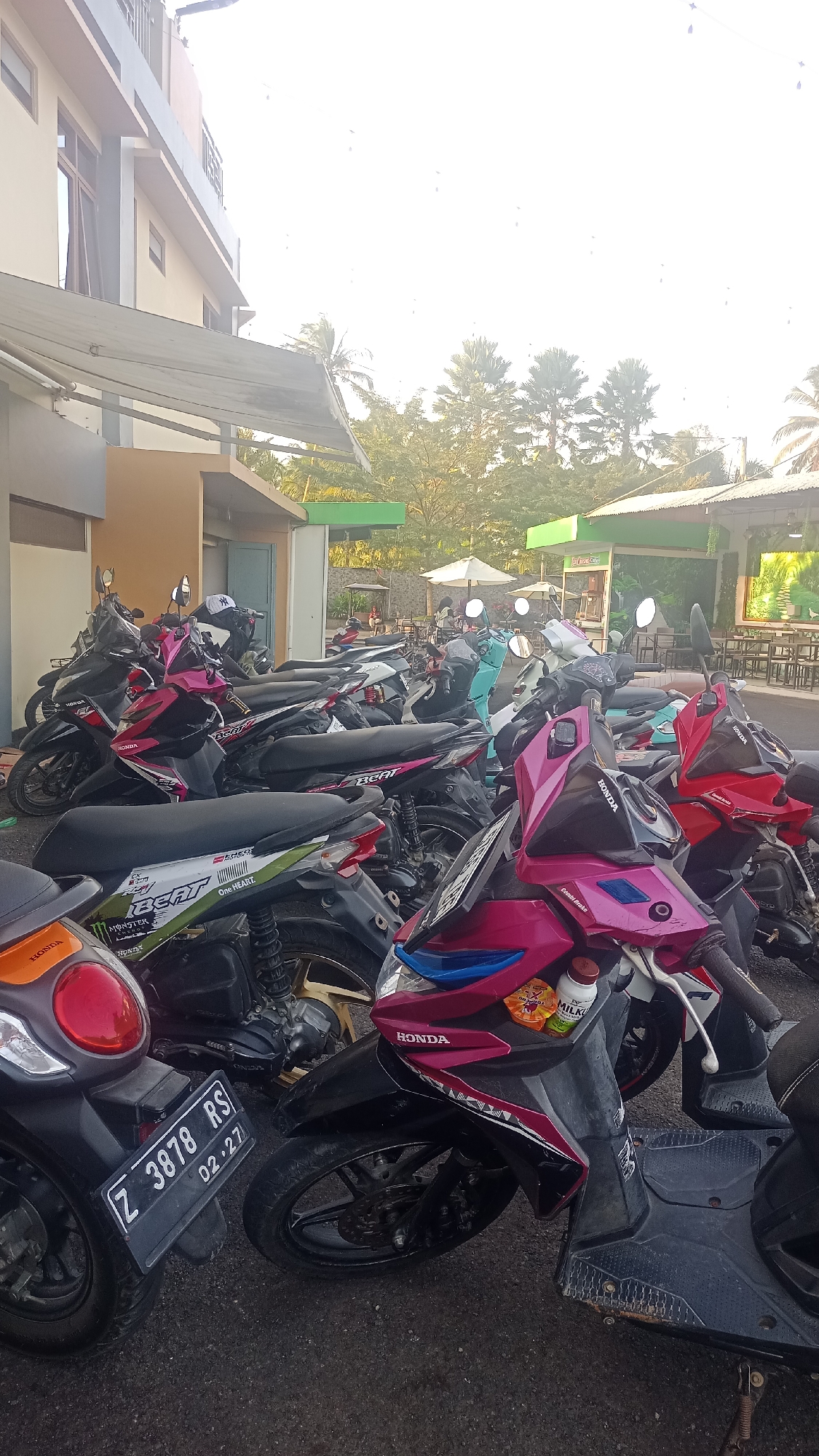 In Pakemitan, Indonesia, a dense arrangement of parked scooters dominates the foreground, with a building and trees visible in the background. The scooters are primarily in shades of black, pink, and red. One scooter in the lower left has a license plate reading "Z 3878 RS 02.27". Several scooters display "Honda" branding. Some scooters have stickers, including one with "MONSTER ENERGY" and "BEAT". A few bottles, possibly drinks, are visible on the handlebars of a pink scooter. The environment appears to be an outdoor parking area adjacent to a building with an overhang. In the background, there are tables and umbrellas, suggesting an outdoor seating area for a cafe or restaurant, surrounded by lush green trees and palm trees. The sky is bright, indicating daylight.