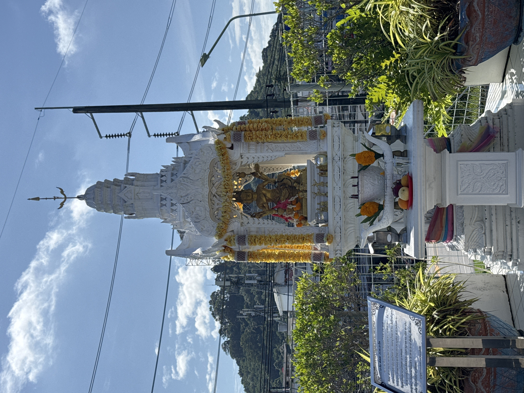 This image depicts an ornate, white spirit house, likely a San Phra Phum, situated outdoors in Wichit, Thailand. The spirit house is elaborately decorated with gold trim, yellow flower garlands, and a golden statue of a deity in the central chamber. Offerings of fruit and incense sticks are visible on a lower tier. The structure is surrounded by lush green foliage in large pots, with a metal fence behind it. In the background, a hill covered in trees and telecommunication towers rises against a bright blue sky with scattered white clouds. A utility pole with power lines runs vertically on the right side of the frame, and a street lamp is visible in the distance. A sign with text in Thai is positioned to the left of the spirit house. The lighting suggests it is daytime.