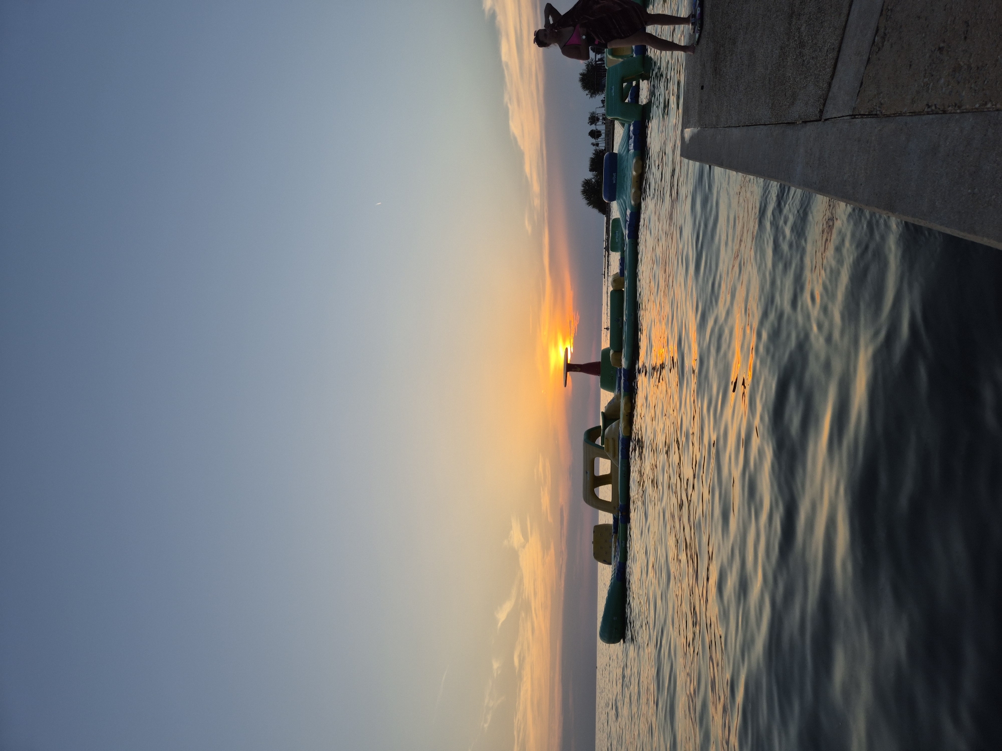 An outdoor, low-angle, wide shot captures a serene sunset over the Adriatic Sea near Katoro, Croatia. The sky displays a gradient of soft blues and oranges, with the sun a bright, glowing orb peeking through the horizon. A long, inflatable water park structure floats on the calm water, its colorful modules casting reflections. In the foreground, a concrete pier runs along the right side of the frame, and on it stands a woman wearing a pink bikini, holding a dark towel over her shoulder. She is facing away from the camera and towards the water. The overall scene evokes a peaceful end to a day by the sea.