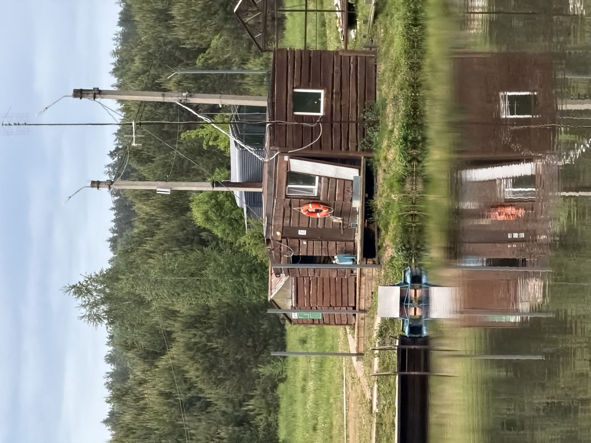 The image shows a two-story wooden structure reflected in water, possibly a lake or river. The structure has windows, a corrugated metal roof, and a life preserver on the exterior. Two utility poles with wires are visible to the left of the structure. The surrounding environment includes trees, grass, and a partially visible metal structure with chairs. The image is likely taken in Prikamskii, Russia.