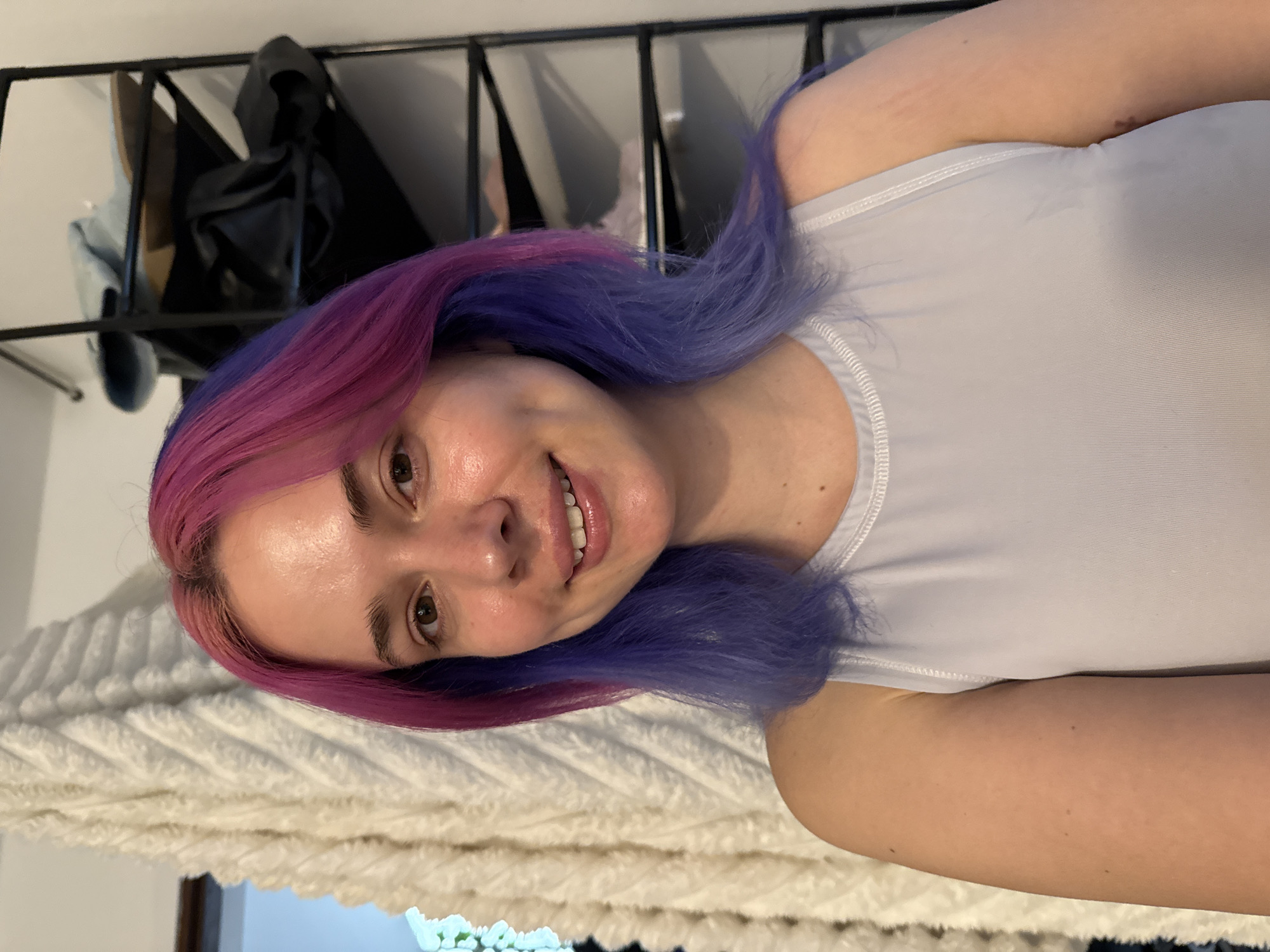 A young woman is depicted in a close-up, chest-up shot, smiling and looking directly at the camera. Her hair is distinctly dyed with vibrant pink on the left side and a complementary blue or purple on the right, falling loosely around her shoulders. She has fair skin, dark eyes, and a cheerful expression, revealing a wide, friendly smile. She is wearing a plain white tank top or athletic-style top, featuring visible stitching along the collar and armholes.
The background is softly blurred but reveals several elements. A white, textured blanket or throw with a wavy, ridged pattern occupies the lower portion of the background, suggesting an indoor setting like a bed or sofa. Above the blanket, a black metal shelving unit or clothing rack is visible, holding various dark fabric items, including what appears to be a black hooded garment, a black bag, and possibly a lighter blue item. The lighting indicates an indoor environment, likely a room such as a bedroom. There are no specific external location cues or visible text within the image. The overall mood conveyed is casual and pleasant.