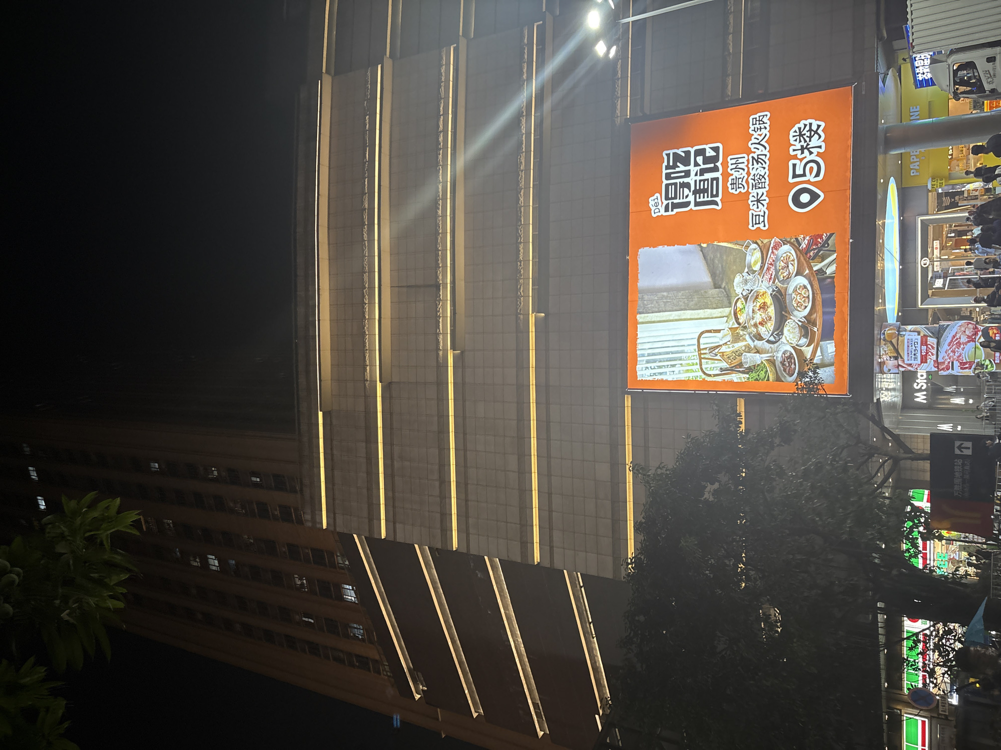 This media file captures a nighttime street scene in the Haizhu District of China. The prominent subject is a large, illuminated billboard advertising "唐记 贵州 豆米酸汤火锅" (Tang Ji Guizhou Dou Mi Sour Soup Hot Pot). The billboard features a vibrant orange background and an appetizing image of a hot pot meal with various ingredients. Text on the billboard indicates the restaurant is located on the "5楼" (5th floor) and mentions "95层" (95th floor), suggesting it's within a tall building.
The billboard is mounted on the side of a modern building with a grid-like facade illuminated by vertical yellow lights. To the left, a darker, older building with visible windows is partially visible. In the foreground, a large, leafy tree obscures some of the view.
The street level below the billboard shows a bustling environment with people walking and shops with illuminated signs. Visible text includes "M Sta", "PAPE NE", and directional signage for "万胜围地铁站" (Wanshengwei Metro Station), confirming the location. The overall atmosphere suggests a busy commercial area at night, likely a shopping district or a food street. The lighting creates a dynamic and urban ambiance. The image appears to be taken from a slightly elevated position, possibly from across the street or a higher floor of an adjacent building.