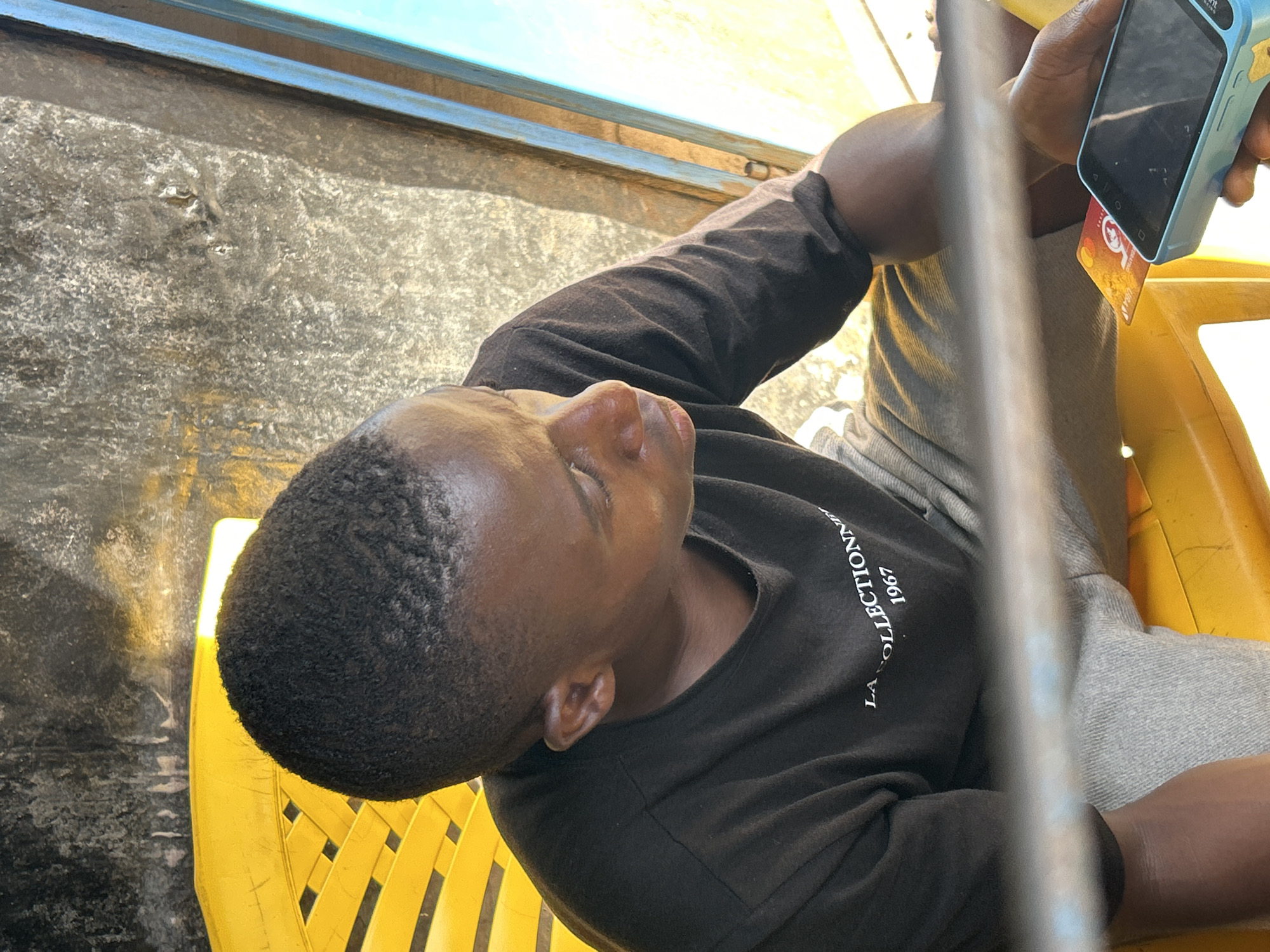 A man with short black hair sits on a yellow plastic chair in an unspecified location in Jos, Nigeria. He is wearing a black long-sleeved shirt with white lettering and gray pants. He is holding a light blue card reader in his right hand, and a gold-colored card is partially inserted into the device. The background includes a textured surface with black and gray tones. A blue and yellow object is partially visible in the upper left corner. A blurred metal pole is positioned in the foreground. The lighting is bright, suggesting an outdoor setting.