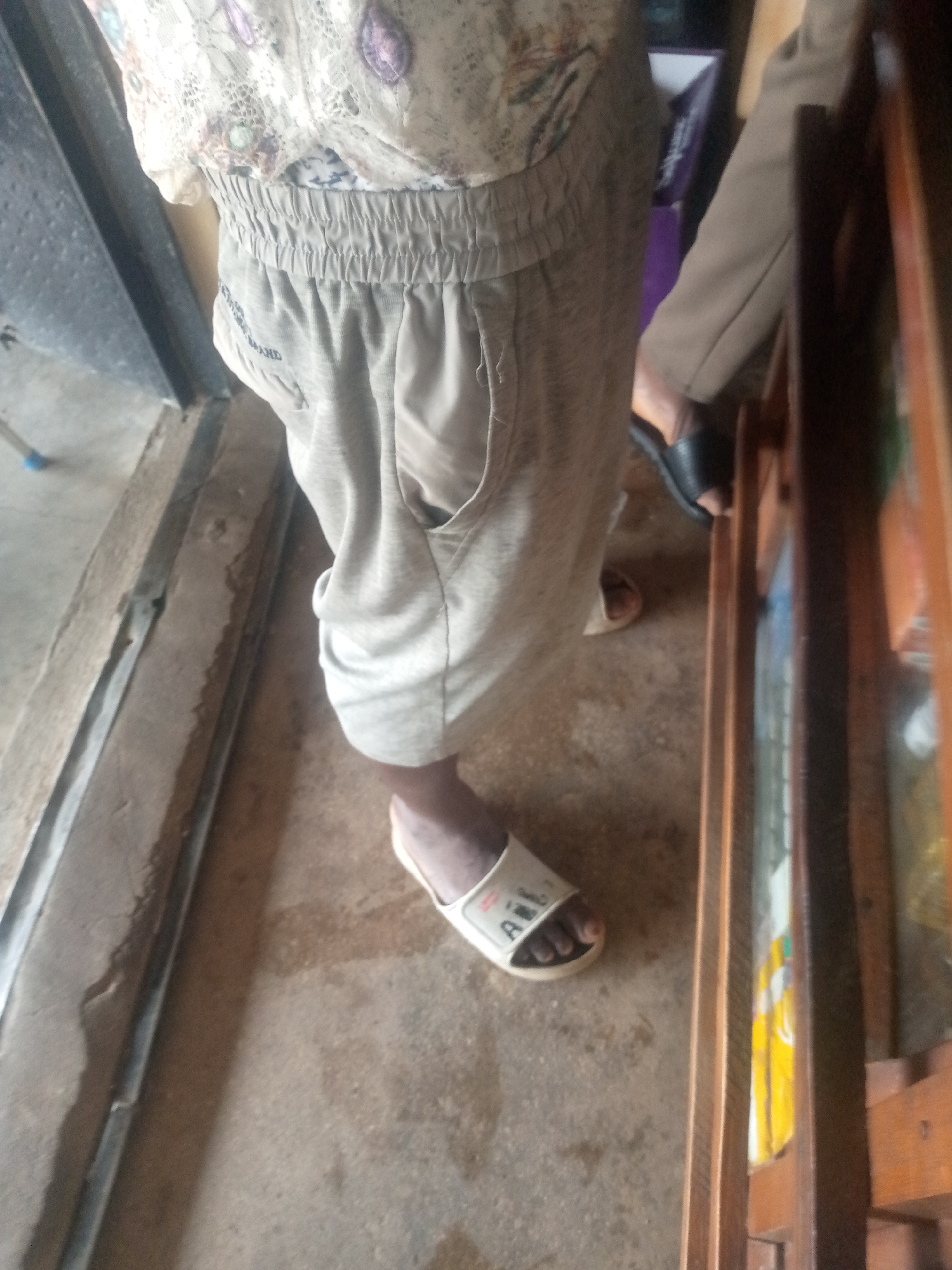 The image shows a person standing in what appears to be a shop or a room with wooden shelves. The person is wearing a light-colored, patterned top and light-colored, loose-fitting pants with an elastic waistband. They are also wearing white sandals with black lettering. The floor is concrete. In the background, there are wooden shelves with some items on them, and part of a doorway is visible. The lighting suggests it is daytime. The scene does not contain any discernible elements that would specifically place it in Serti, Nigeria.