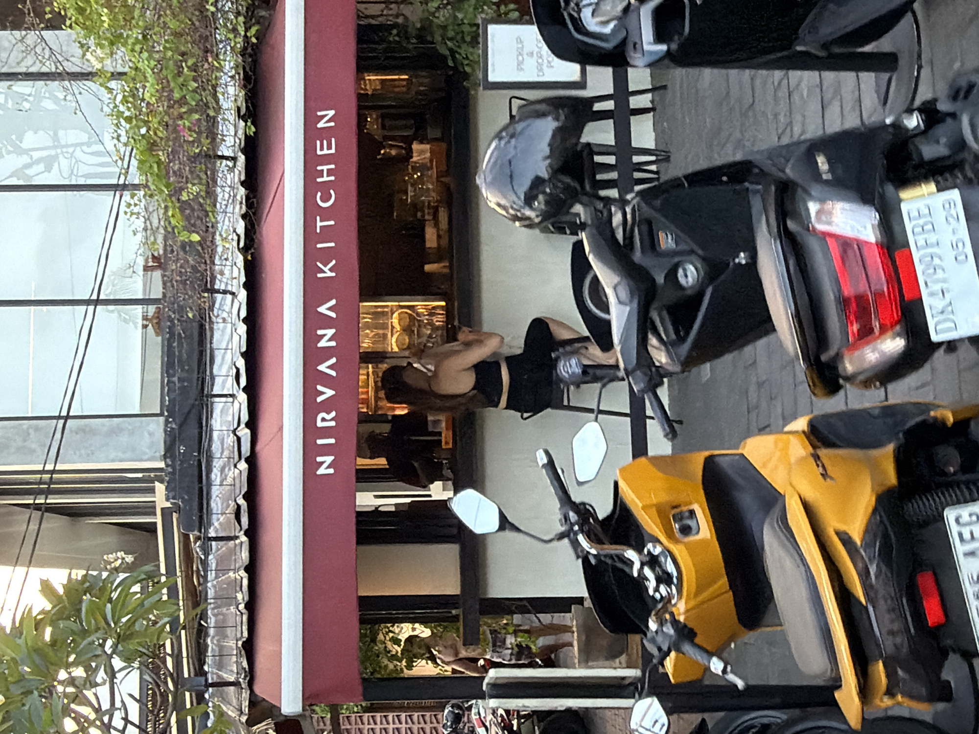 The image depicts the storefront of "NIRVANA KITCHEN" in North Kuta, Indonesia. A red awning with white lettering marks the establishment. In front of the eatery, several parked scooters are visible, with a yellow scooter prominently featured in the foreground. A woman with dark hair is seated at a counter inside the kitchen, facing away from the camera and holding a mobile phone. To her left, a shirtless man wearing patterned shorts stands near a potted plant. Behind the counter, a person, possibly an employee, is visible in silhouette. The exterior features a tiled facade and a row of chairs outside. A white sign with black text indicating "PICKUP & DROP-OFF" is also visible. The scene appears to be outdoors, with natural light illuminating the area.