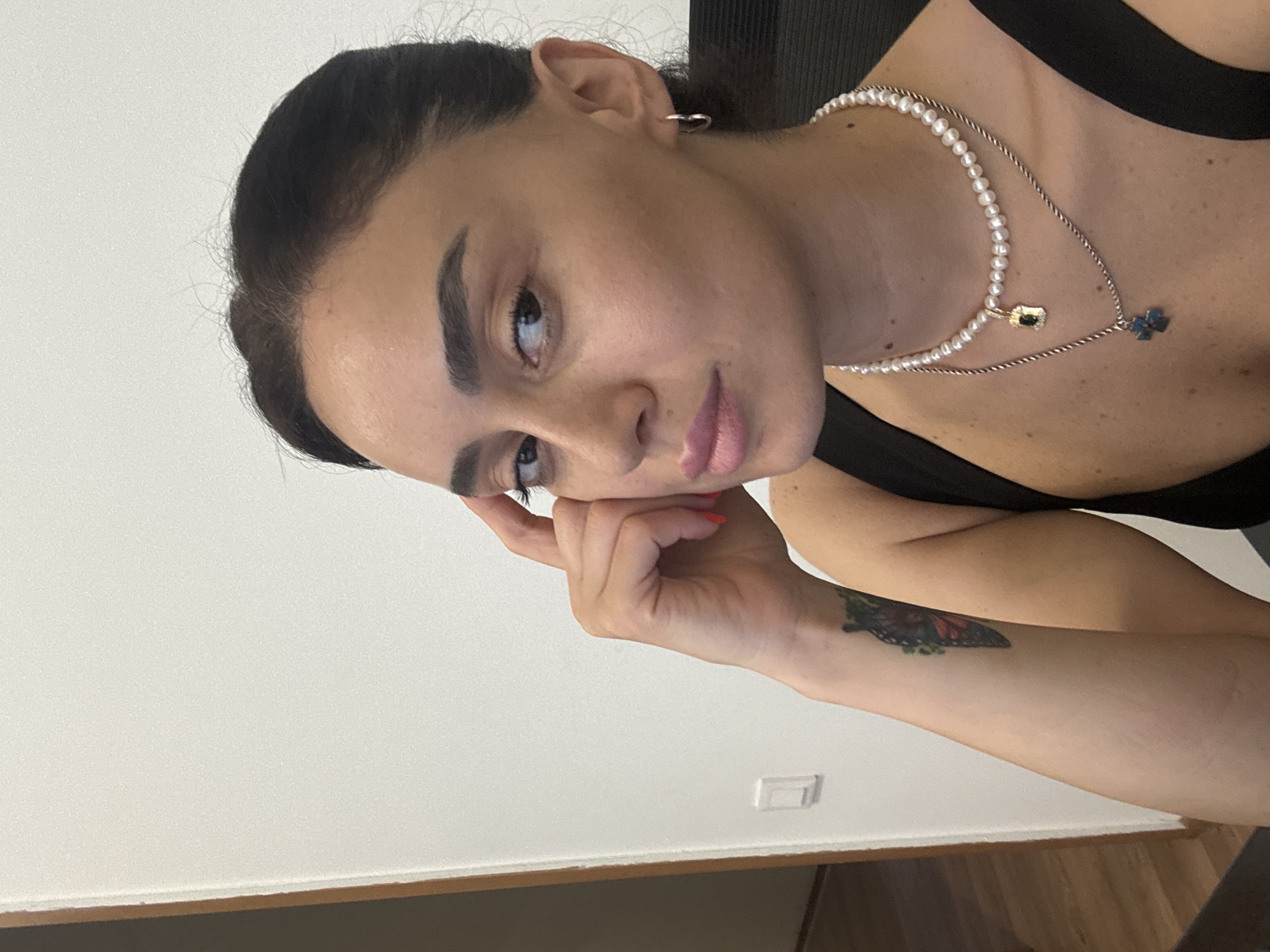 A woman of Middle Eastern descent is pictured indoors, seated, with her right hand resting on her temple. She has dark hair pulled back, prominent eyebrows, and is wearing a black tank top, a pearl necklace with a gold pendant, and a silver chain with a blue cross pendant. She is also wearing small silver hoop earrings. A tattoo of a butterfly is visible on her left forearm. The background consists of a plain white wall, a doorway with a wooden frame to the left, and a glimpse of a dark chair behind her. An electrical outlet is visible on the white wall to her left. The overall lighting suggests an indoor environment, possibly an office or residential space, with no specific identifying markers of Tbilisi, Georgia.