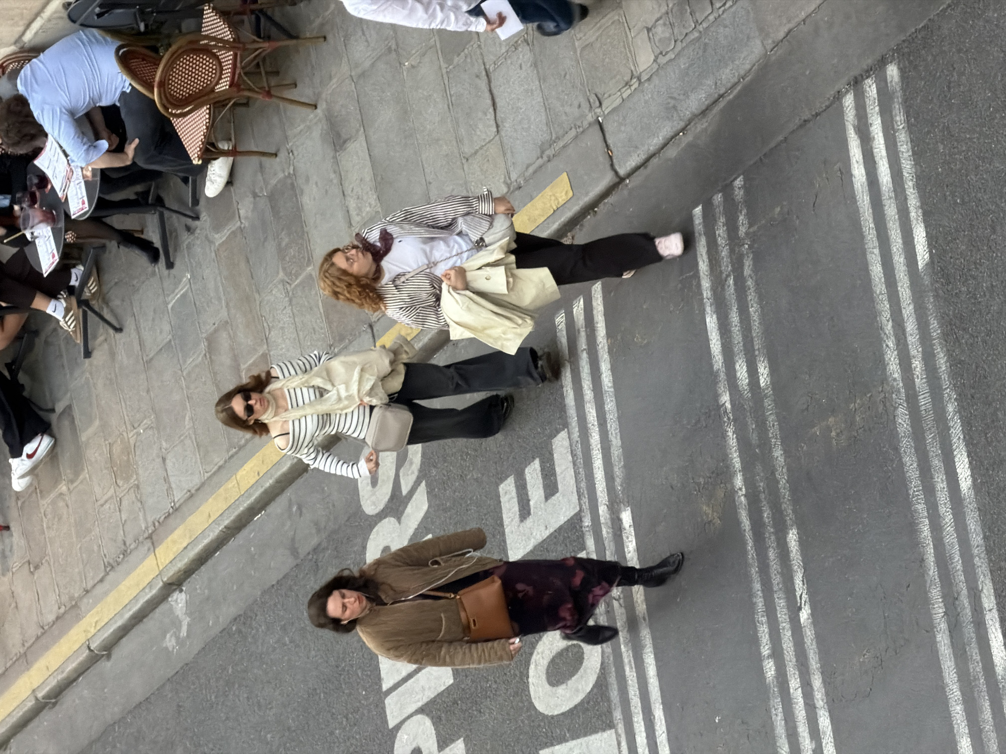 The image captures a high-angle, tilted view of an urban street scene, likely in Paris, France, from an overhead perspective looking downwards. The scene is divided by a curb and a yellow line separating a paved sidewalk from an asphalt street.
On the left, an outdoor cafe terrace is visible on the paved sidewalk. Two individuals are seated at a small, dark round table with brown wicker chairs. One person, wearing a light blue shirt, leans forward over the table. Another person's legs are visible, wearing dark pants and white sneakers with prominent Nike socks. Menus and drinks, including a glass with a reddish liquid, are on the table.
On the right side of the image, the asphalt street and the edge of the sidewalk are populated by three women standing or walking.
- One woman with auburn, curly hair wears a striped long-sleeve jacket, a white t-shirt, black pants, and light pink shoes. She carries a cream-colored tote bag and a smaller light-colored shoulder bag.
- Another woman, with dark hair, wears a striped long-sleeve top, sunglasses, a light-colored scarf, black pants, and white Nike sneakers. She carries a small, light-colored cross-body bag.
- A third woman, with dark hair, wears a brown quilted jacket, a dark patterned skirt, and black boots. She carries a brown leather cross-body bag and has earbuds in.
The asphalt street features prominent parallel white stripes, possibly shadows cast by unseen overhead structures or faded road markings. Faint, large white letters are visible on the street, clearly spelling "OLE" or "EOL" (depending on rotation), suggesting part of a larger word or sign like "ECOLE" or "VELO". The lighting suggests it is daytime.