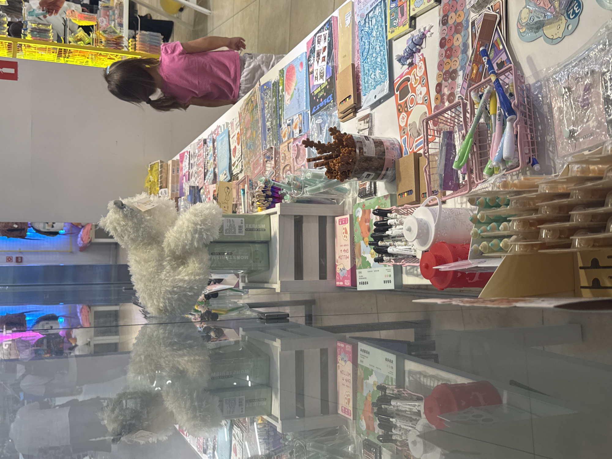 A young girl with brown hair in a pink t-shirt and gray shorts stands in front of a table displaying various stationery items. The table is laden with colorful stickers, notebooks, pens, and other craft supplies. Behind the girl, a yellow retail display rack is visible, filled with more merchandise. The scene appears to be set in a store, likely a stationery or gift shop. The lighting is bright, suggesting an indoor environment. The overall impression is one of a retail setting with a focus on creative and decorative items. The reflection in the glass in the foreground indicates that the viewer is looking into the store from the outside, possibly through a window or display case.