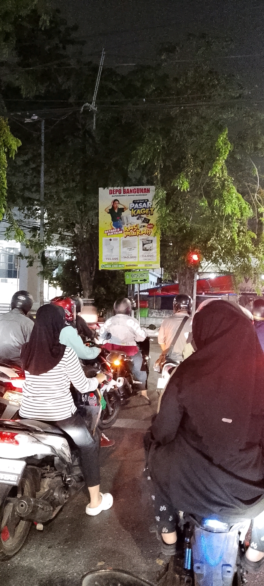 The image captures a nighttime scene on a street, likely in Bekasi, Indonesia, showing a queue of motorcyclists waiting at a traffic light. A large banner hangs from trees on the right side of the street, advertising "DEPO BANGUNAN SUPERMARKET BAHAN BANGUNAN PASAR KAGET" (Depo Bangunan Building Materials Supermarket Surprise Market). The banner features a woman with her hands on her head and text promoting "KASTI MURAH" (Certainly Cheap). Prices for various items are listed below: 755,000, 149,000, and 114,000. A phone number, 0821 2339 9979, is also visible.
In the foreground, several individuals are on motorcycles. One person in the immediate foreground, wearing a black hijab and a black abaya, is seated on a motorcycle facing away from the camera. To their left, another person wearing a striped long-sleeved shirt and black pants, also on a motorcycle, is visible. A water bottle is placed in a holder on their motorcycle. Other motorcyclists and their passengers, mostly wearing helmets, are also part of the traffic. The environment is characterized by trees lining the street and the glow of streetlights and motorcycle lights, indicating it is nighttime. The road surface appears wet, suggesting recent rain. The overall atmosphere suggests a typical urban traffic situation at night in Indonesia.