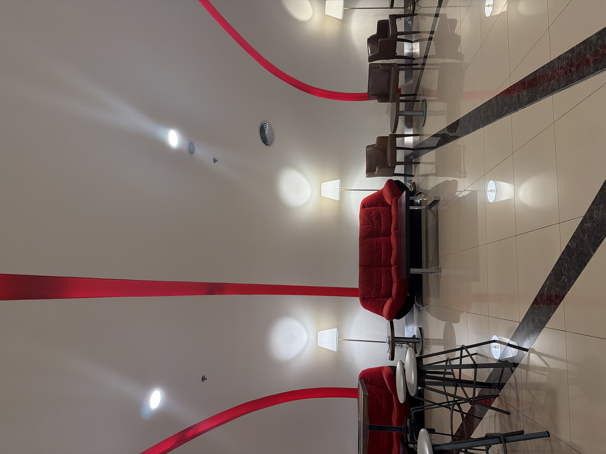 An overhead shot looking down on a room with red couches and brown chairs in Sofia, Bulgaria. The scene is a lobby or waiting area with cream-colored tiled floors, black decorative floor inlays, and white walls. Red curved stripes run along the walls near the ceiling. Two red couches sit at the bottom of the frame, with white side tables and floor lamps. Adjacent to the couches are several brown chairs with small tables, all arranged in a line. The lights from the lamps illuminate the scene, and there are recessed lights in the ceiling. No people are visible.