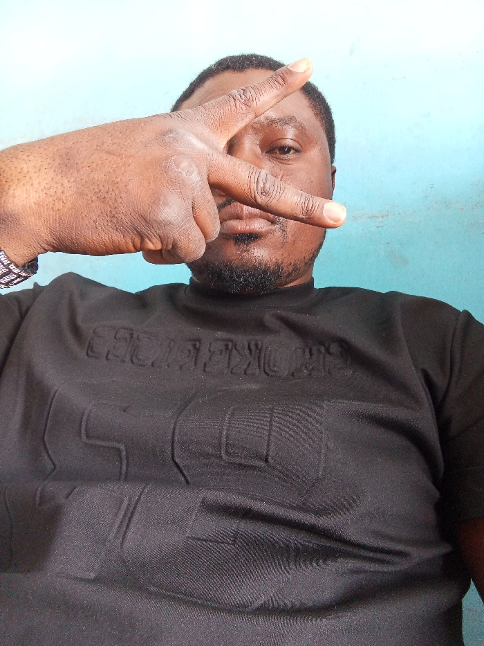 The image displays an adult male, dark-skinned, with short dark hair and a trimmed beard and mustache. He is positioned centrally, facing the viewer with a neutral expression. His left hand is raised, fingers forming a 'V' gesture with the index and middle fingers extended, partially obscuring his left eye and forehead. He wears a black, short-sleeved, high-collared shirt featuring embossed text on the chest, including words resembling "STOKES" and a large numerical design, possibly "59". A multi-colored, beaded bracelet is visible on his left wrist. The background is a plain, light blue wall. The specific location within the city of Jos, Nigeria, is not discernible from the visual information provided.