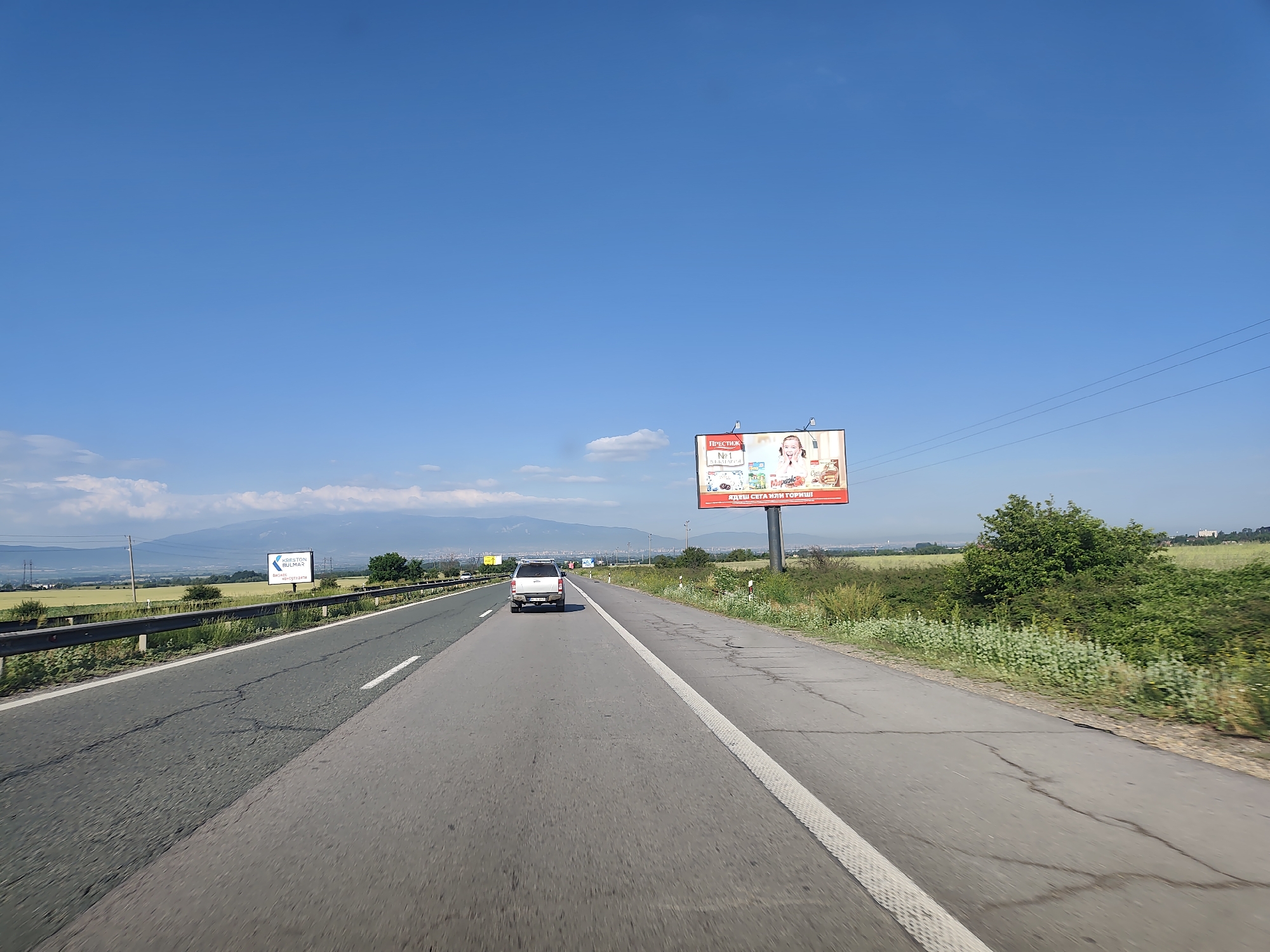The image depicts a highway scene in Yana, Bulgaria, under a clear blue sky. A white SUV is driving on the right lane of the highway, with a guardrail separating the highway from a grassy embankment on the left. On the right side of the highway, a billboard displays an advertisement for "PRESTIGE" products. In the distance, mountains are visible, along with agricultural fields and some trees. Another smaller billboard is seen in the far distance on the left side of the highway. The road surface shows signs of wear with visible cracks.