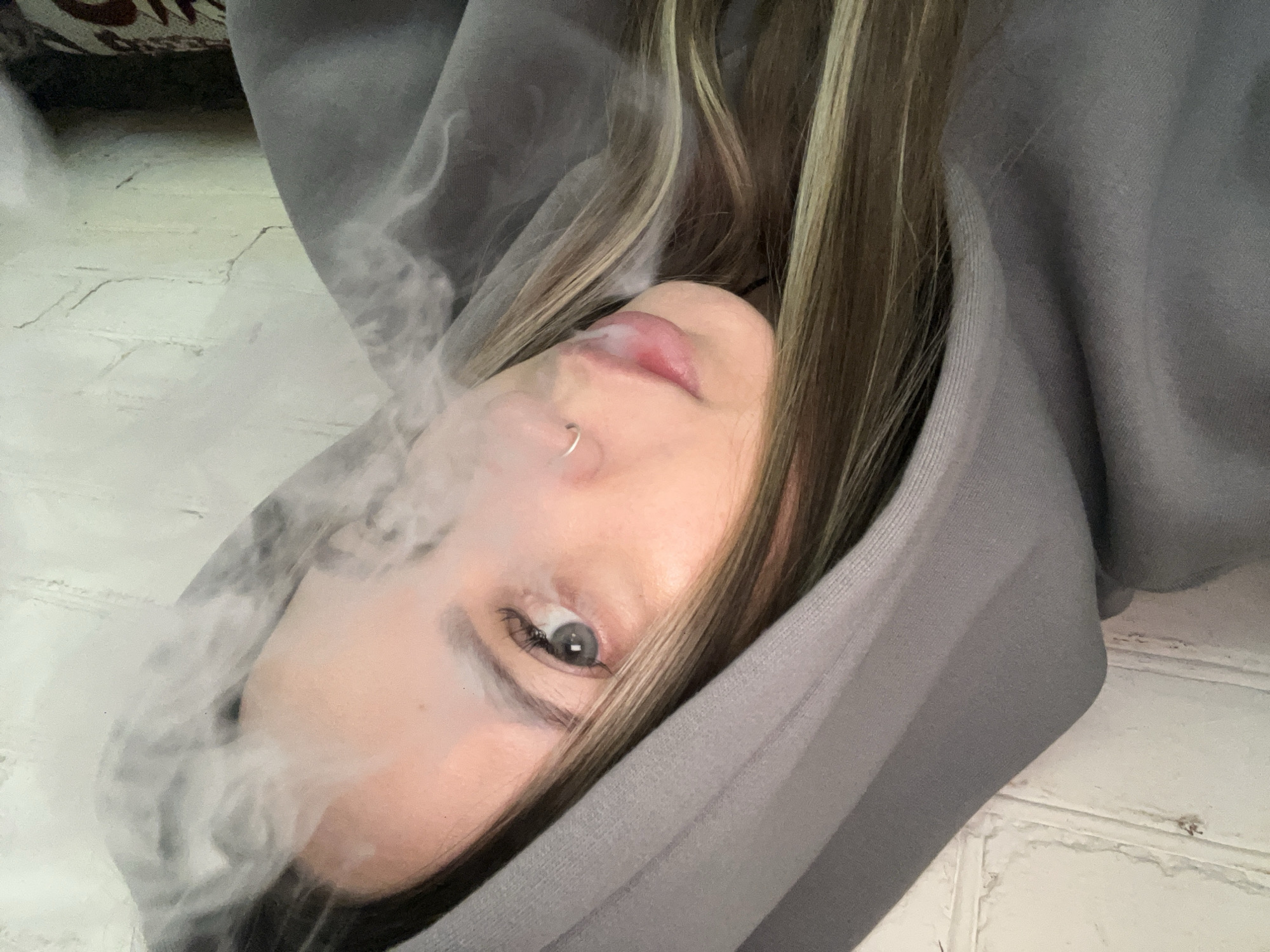 A close-up, horizontally oriented image captures a young person, likely female, exhaling a large cloud of white smoke or vapor from their mouth. The subject is wearing a grey hooded sweatshirt, with the hood framing their face. Their long, brown hair with blonde highlights cascades around their head. A small silver hoop piercing adorns their nose. One light-colored eye, possibly blue or green, is visible and gazes directly at the camera, while the pink lips are slightly parted as the smoke emerges. The dense vapor obscures the left side of the person's face and parts of the background. The setting appears to be indoors, with a light-colored, textured surface (possibly a painted brick wall or floor) visible in the background, along with a darker, out-of-focus object that could be furniture in the upper left corner. The activity is focused on the exhalation of smoke. No visible text is present in the image. The location context is Novopokrovskaya, Russia.
