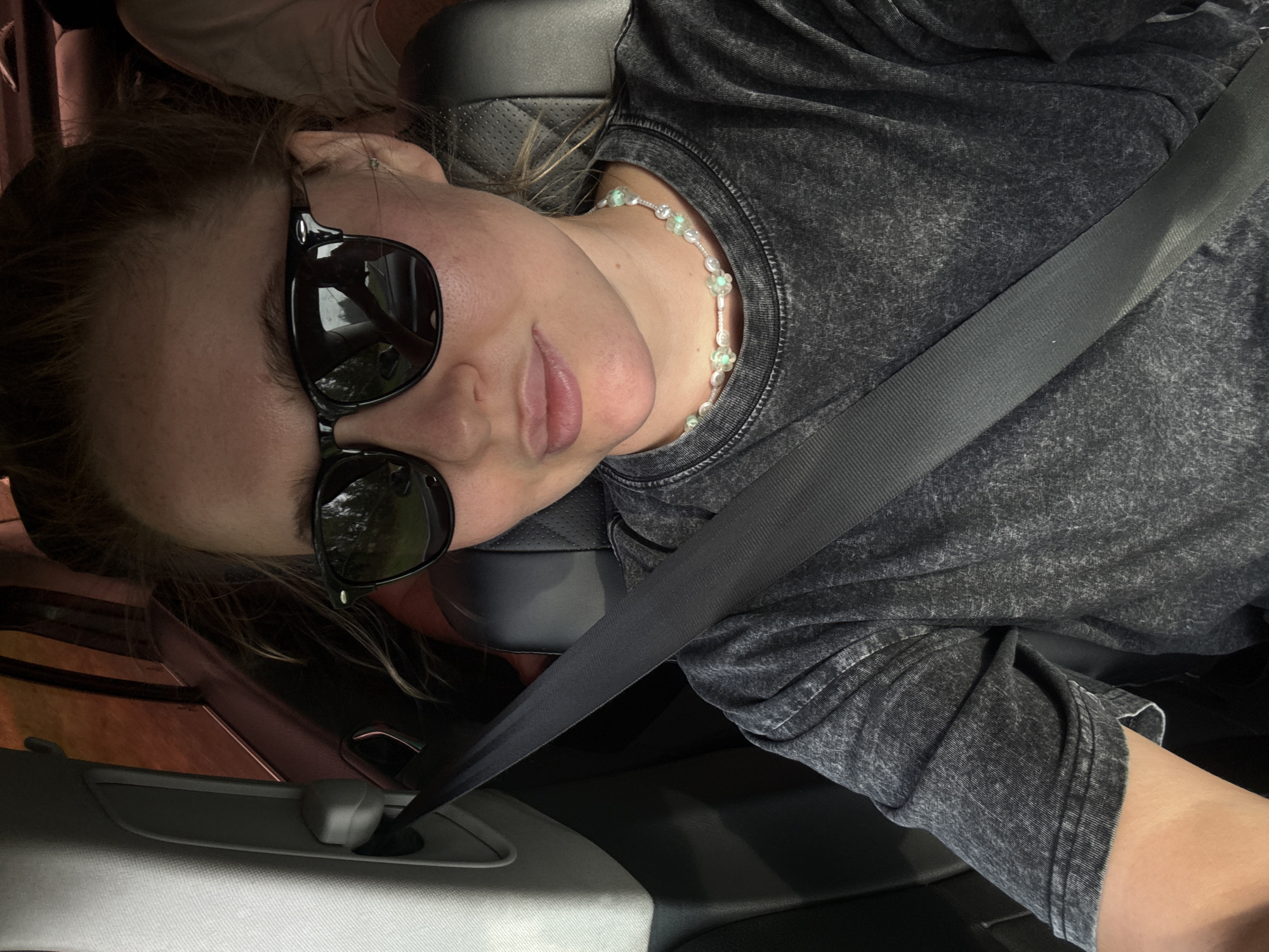A young woman wearing sunglasses and a dark grey t-shirt sits in the passenger seat of a car. She is wearing a beaded necklace. A seatbelt is fastened across her chest. The interior of the car, including the grey plastic trim and black leather seat, is visible. The background shows a blurred view of trees and a road, indicating the car is in motion. The image was taken in Nylga, Russia.