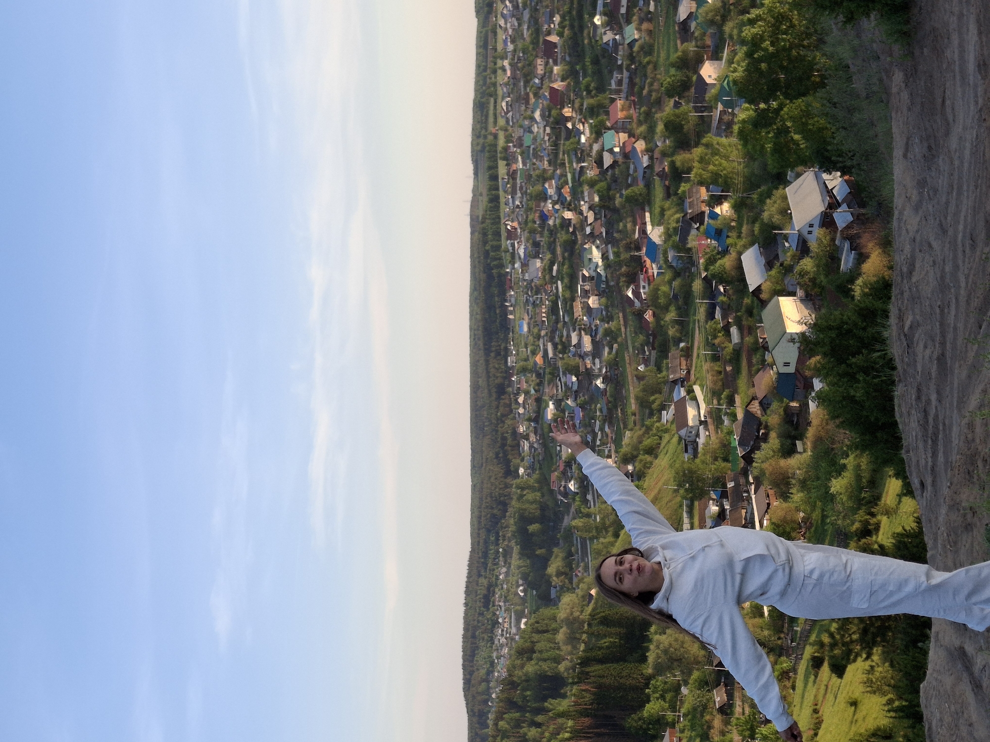 A full shot depicts a person with their right arm raised, standing on a rocky edge, overlooking a sprawling cityscape. The cityscape consists of many residential buildings, some with brightly colored roofs. Between the city and the sky is a dense, dark forest. The person is wearing white and is centered in the foreground, with the sky and city filling the background. The location is Ольховка, Russia.