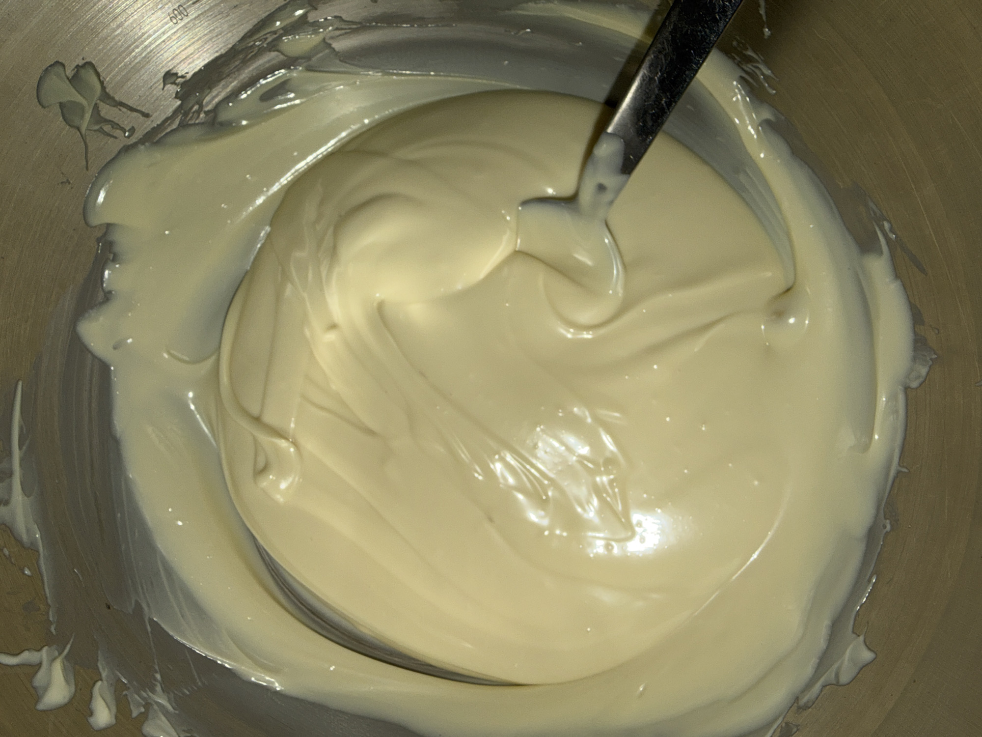 A close-up overhead shot depicts a silver metal mixing bowl filled with a large quantity of smooth, light yellow or off-white frosting or cream. A metal spoon is partially submerged in the center of the bowl, with its handle extending upwards. The frosting/cream is glossy and has swirling patterns, suggesting it has recently been stirred. Some of the mixture adheres to the sides of the bowl, indicating previous mixing or spreading. The environment lacks recognizable markers of Strasbourg, France.