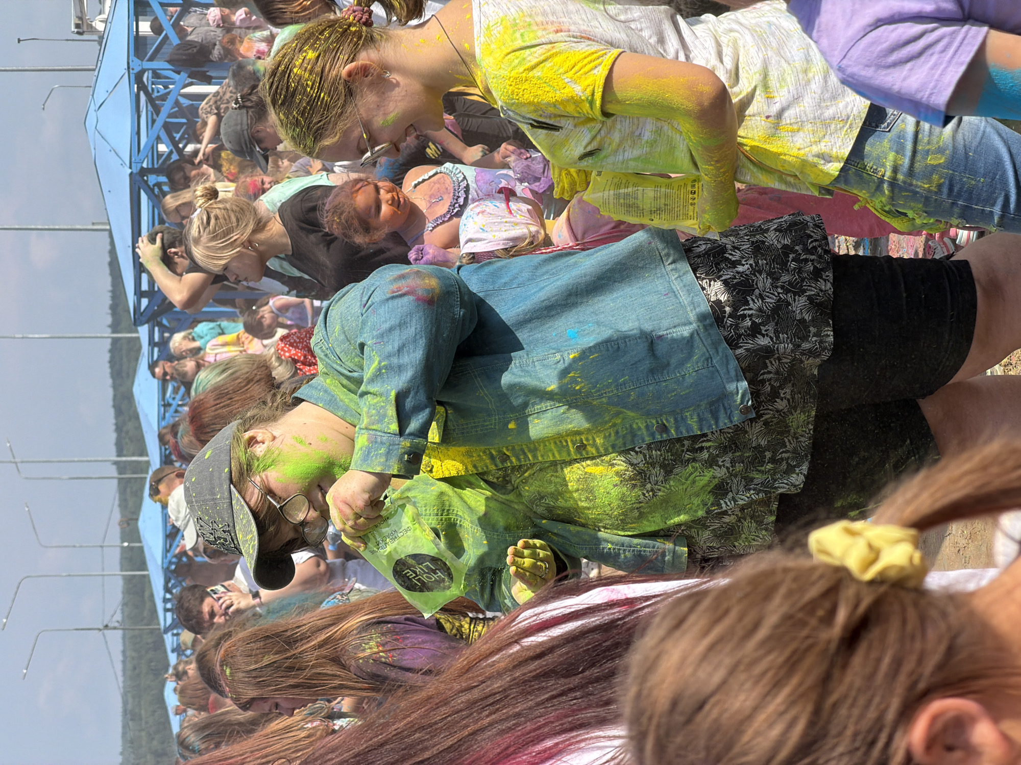The image depicts a crowded outdoor festival, likely a color run or a similar event, occurring on a sunny day in Chaikovsky, Russia. The foreground features several young people covered in bright green and yellow powder, indicative of the Holi festival. One individual in the center wears a denim jacket and a baseball cap, holding a green packet of powder. To the right, another person in a white t-shirt and jeans is also adorned with yellow powder. In the background, numerous other attendees are visible, some with colored powder on their clothing and hair. Structures with blue roofing and tents suggest a designated festival area. The overall atmosphere appears festive and energetic.