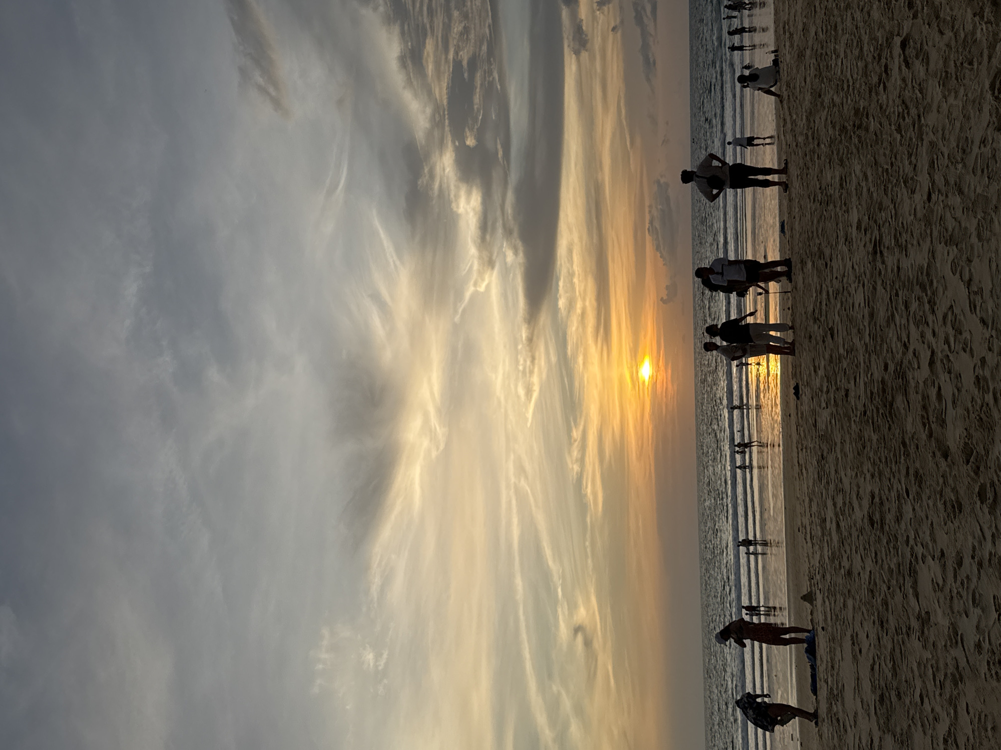 The image depicts a sunset scene at Karon Beach in Karon, Thailand. Silhouetted figures of people are scattered across the sandy beach and shallow water. Some individuals are walking, while others are standing or sitting. The sun, partially obscured by clouds, is low on the horizon, casting a warm orange glow across the sky and reflecting on the wet sand. The ocean stretches out towards the horizon, with gentle waves breaking on the shore. The sky is filled with a mix of soft grey and wispy clouds illuminated by the setting sun. The overall atmosphere is serene and tranquil, capturing a typical evening on a popular Thai beach.