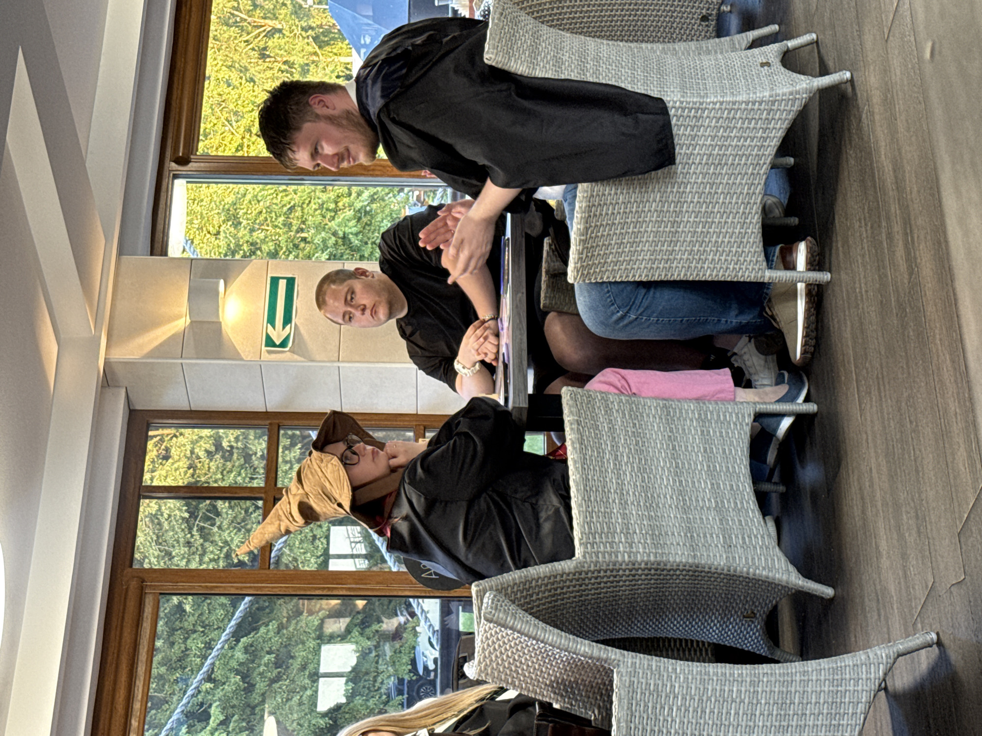 Three individuals, two males and one person appearing to be female wearing a pointed hat and glasses, are seated at a table. The person in the hat is wearing a dark robe and pink pants. The male to their left is wearing a black t-shirt and jeans, and the male to their right is also wearing a dark robe. They are in an indoor setting with large windows overlooking trees. The room has a modern design with tiled walls and a tiled floor. A green directional arrow sign is visible on the wall. The image does not contain any elements that would suggest a specific location within Veliky Novgorod, Russia.