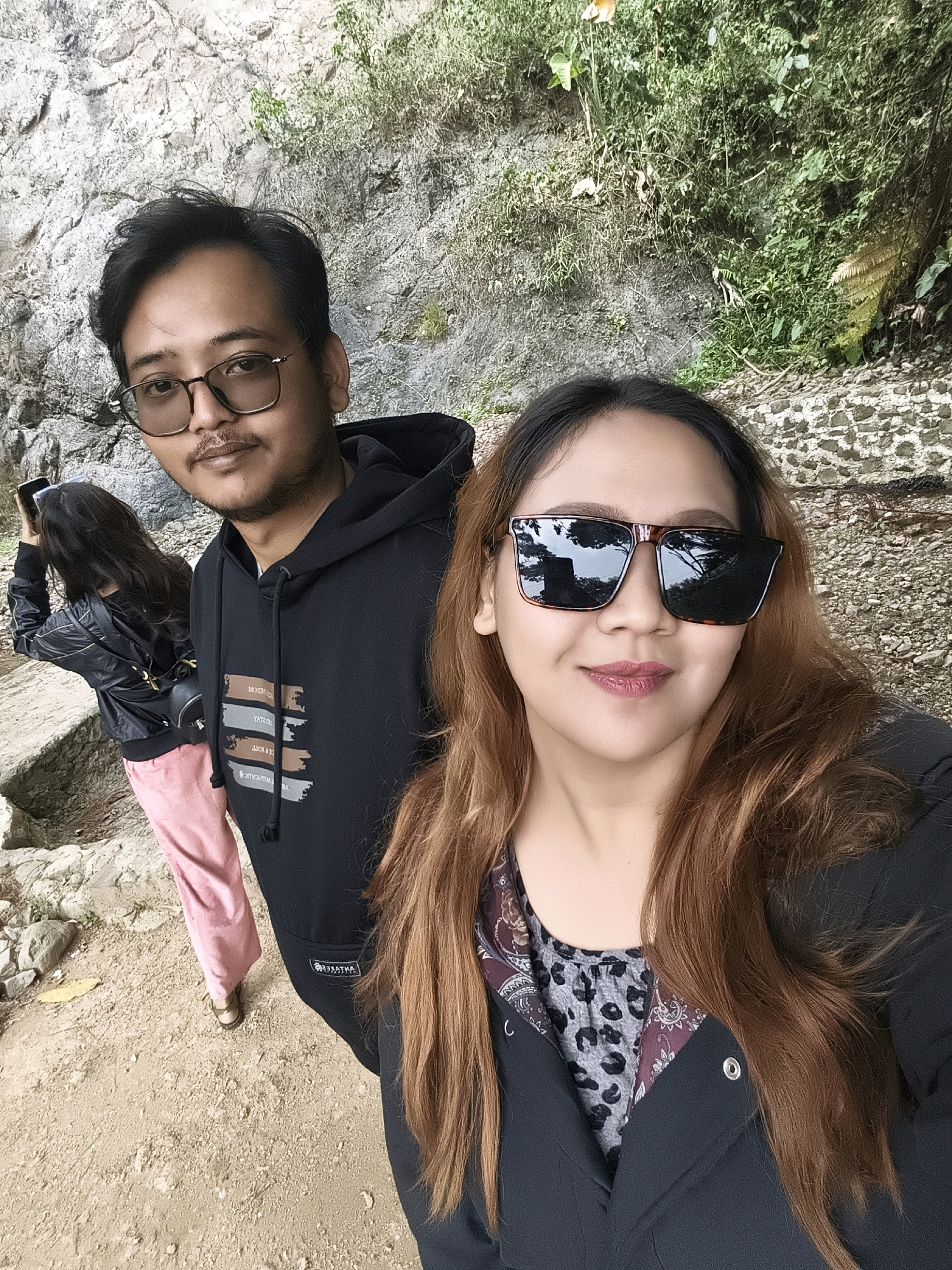 A medium shot captures three individuals outdoors, with two in the foreground and one in the mid-ground. The primary subject is a woman in the foreground, facing the camera and smiling, wearing large, dark sunglasses, a black jacket, and a patterned top. To her left, a man with glasses, a mustache, and a goatee is also facing the camera, wearing a black hoodie. Behind them and to the left, another person, seen from the back, is holding a phone to their ear and wearing a black jacket and pink pants. The environment is a rocky and vegetated area with a dirt path in the foreground. The scene appears to be a selfie, likely taken during an outing or hike.