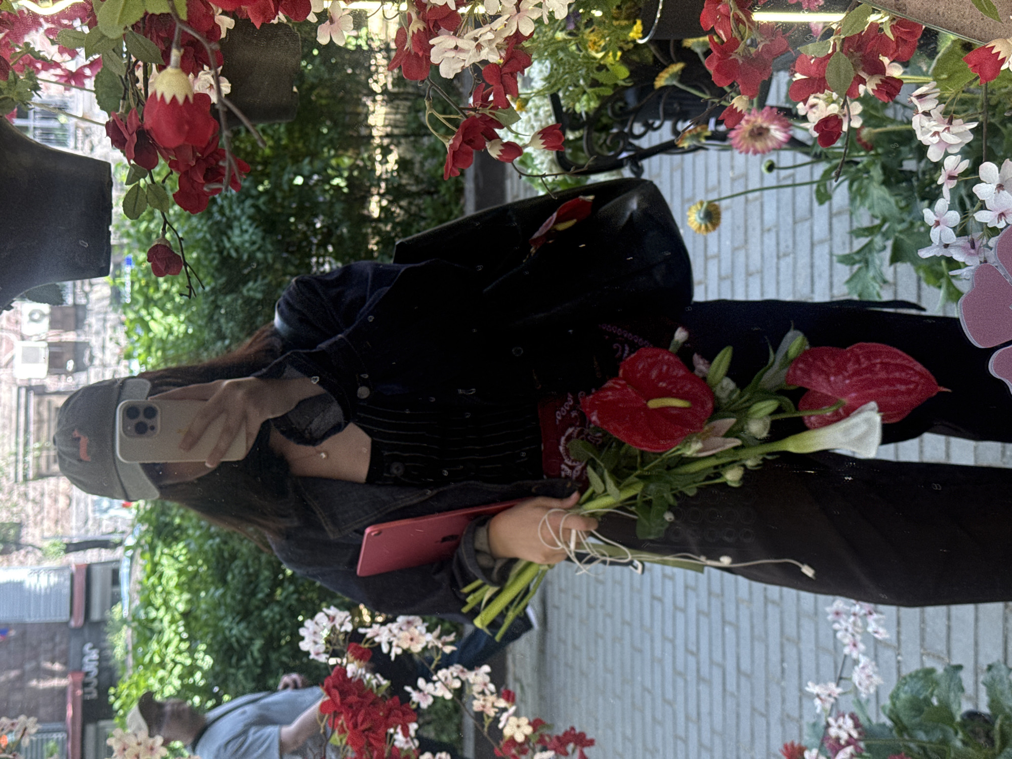 A person stands in front of a mirrored surface in Yerevan, Armenia, holding a bouquet of red anthurium flowers and a red tablet. The person wears a dark jacket, a baseball cap, and a face mask. The mirrored surface reflects the surrounding environment, including a pedestrian wearing a blue shirt and green trees. Decorations of red and white flowers frame the upper part of the mirrored surface. The reflection shows urban buildings and a tiled sidewalk. The person appears to be taking a selfie using a smartphone.