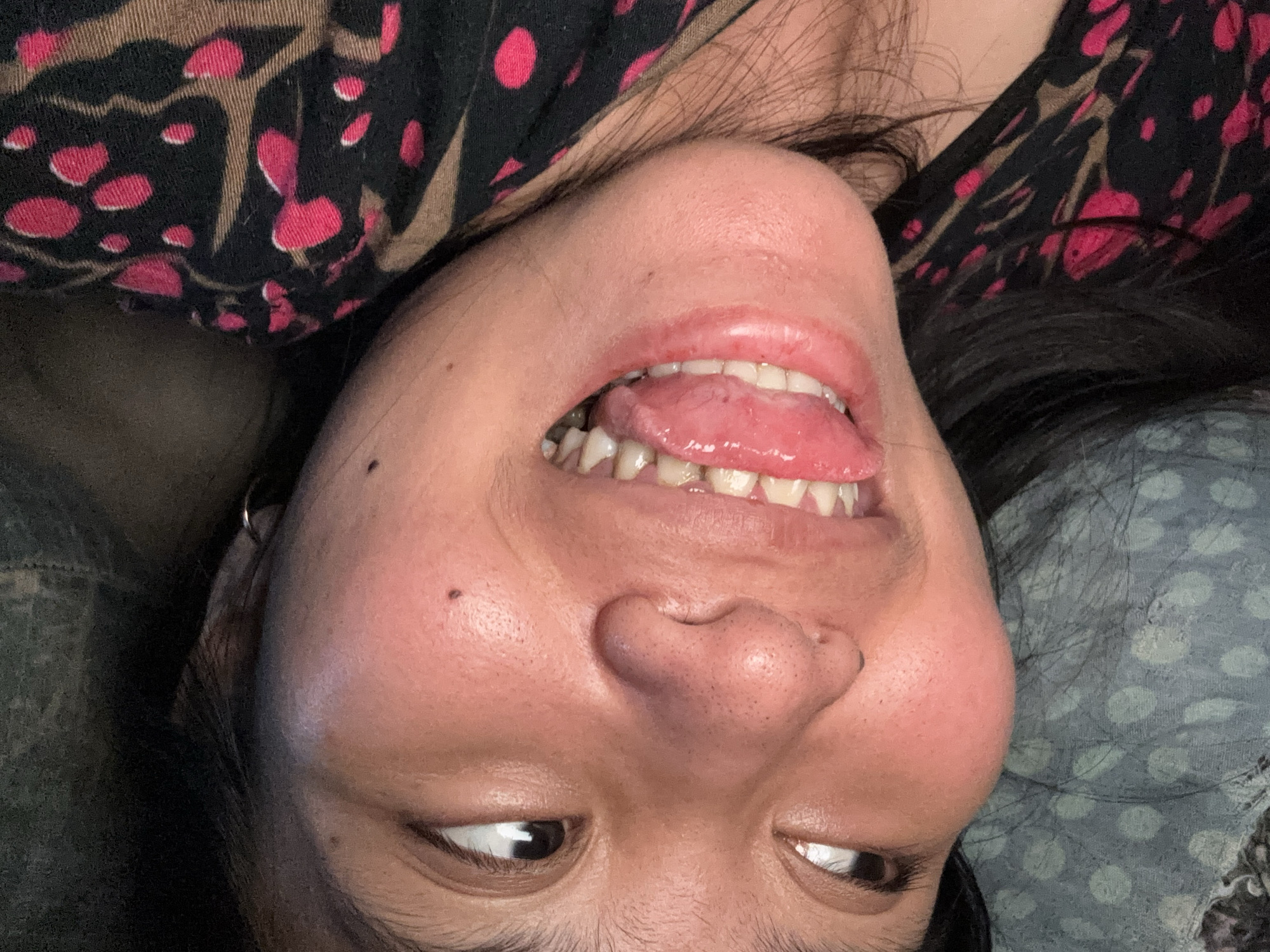 The image shows a close-up, sideways portrait of a person's face. The person has dark hair and is wearing a black garment with a pink and brown pattern. Their mouth is open, revealing teeth and a pink tongue. The background is dark and appears to be a soft surface, possibly fabric or bedding. The image is a casual, self-taken photo, likely indoors. The location in East Jakarta, Indonesia, is not discernible from the image itself.