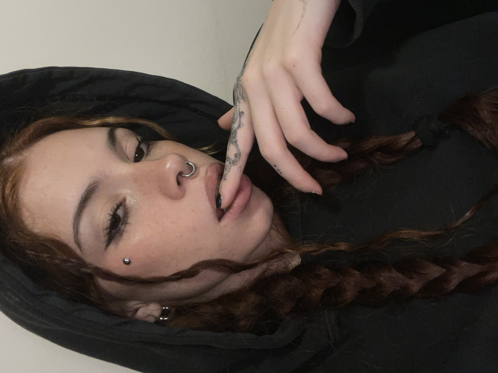 A young woman lies on her back, positioned against a white or light-colored wall. She is wearing a black hooded sweatshirt. The hood frames the side of her face. She has reddish-brown hair, styled in braids.
She has fair skin with freckles, and several piercings. A ring is visible in her nostril, a stud piercing on her cheek, and an earring in her ear. She is wearing eyeliner. Her mouth is slightly open and her finger is positioned near her lips. The tattoo is visible on her hand.
The location cannot be determined from the image. The image does not have the information to place it in Buenos Aires, Argentina.