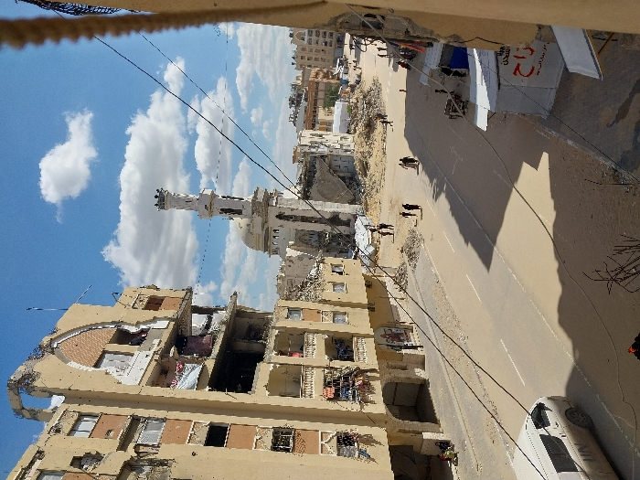 This image depicts a street scene in Khan Yunis, Palestinian Territory, characterized by extensive urban destruction. On the left, a multi-story, light-colored building stands severely damaged, with multiple floors exposed to the elements, large sections of its facade missing, and rebar visible. Directly below it, a white van with "UN" clearly marked on its side is visible on the sandy road. The street, partially covered in sand and debris, extends into the midground where several individuals, dressed in dark clothing, walk. In the background, a mosque with a distinctive minaret and dome shows significant structural damage and partial collapse, surrounded by piles of rubble. Other partially destroyed buildings and debris fields are visible throughout the background. The sky is bright blue with scattered white cumulus clouds. On the right side of the street, makeshift shelters or stalls with white tarps are present, and electrical wires traverse the upper portion of the frame. Several small groups of people are scattered across the scene, navigating the street and rubble-strewn areas.