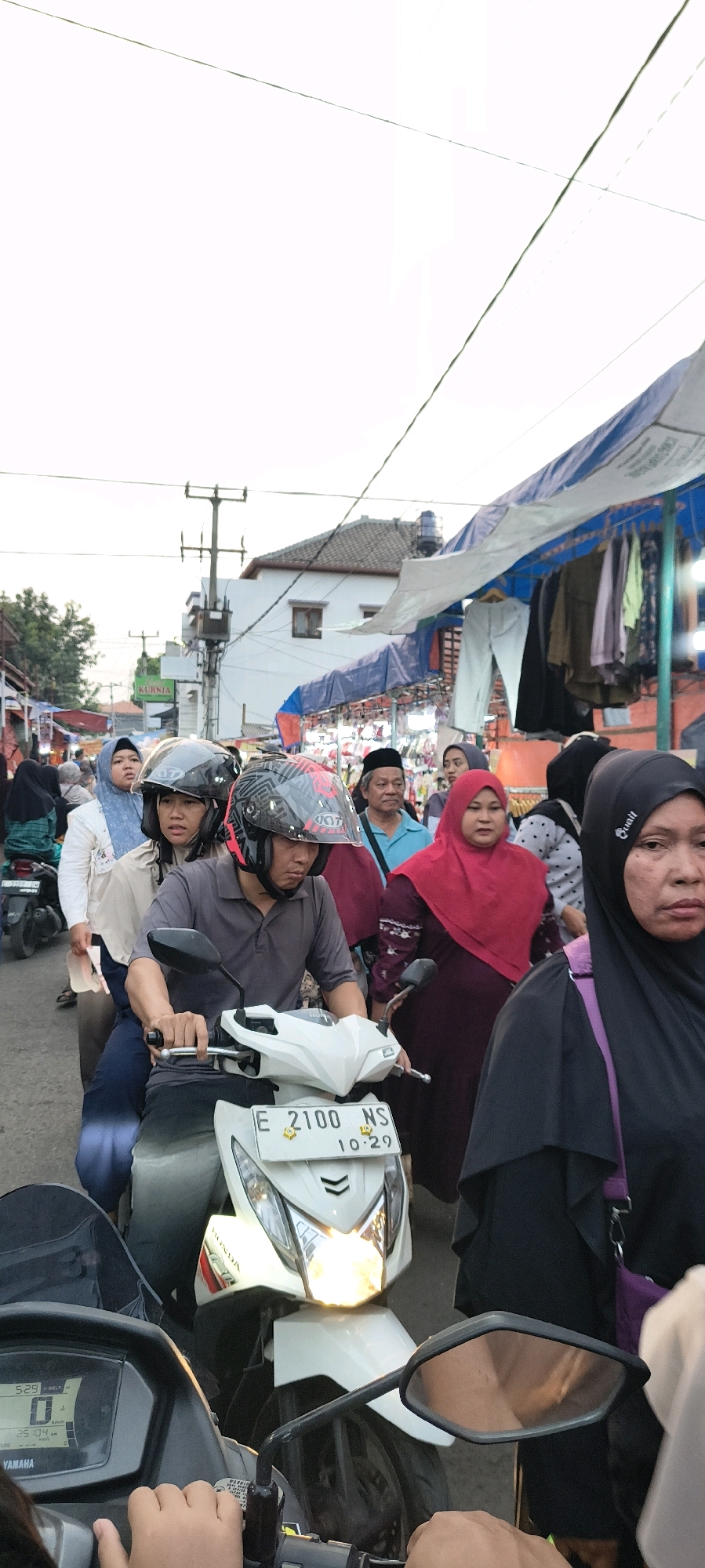A low-angle, vertical, full-shot image captures a street scene in Trusmi Wetan, Indonesia, with a focus on a white scooter in the foreground. The scooter, bearing license plate "E 2100 NS" and the date "10-29," has its headlights illuminated. A person wearing a grey polo shirt and a black helmet is operating the scooter, with another person in a light-colored hijab and a dark helmet riding as a passenger.
Behind them, a crowded street market unfolds. Several people are visible, many wearing hijabs. A woman in a red hijab and a dark patterned dress stands out, as does a woman in a black hijab on the far right of the frame. Market stalls are set up under blue and white awnings, displaying clothing and other goods. In the background, buildings with tiled roofs and utility poles suggest a residential or commercial area. The sky is overcast. The image appears to have been taken during daylight.