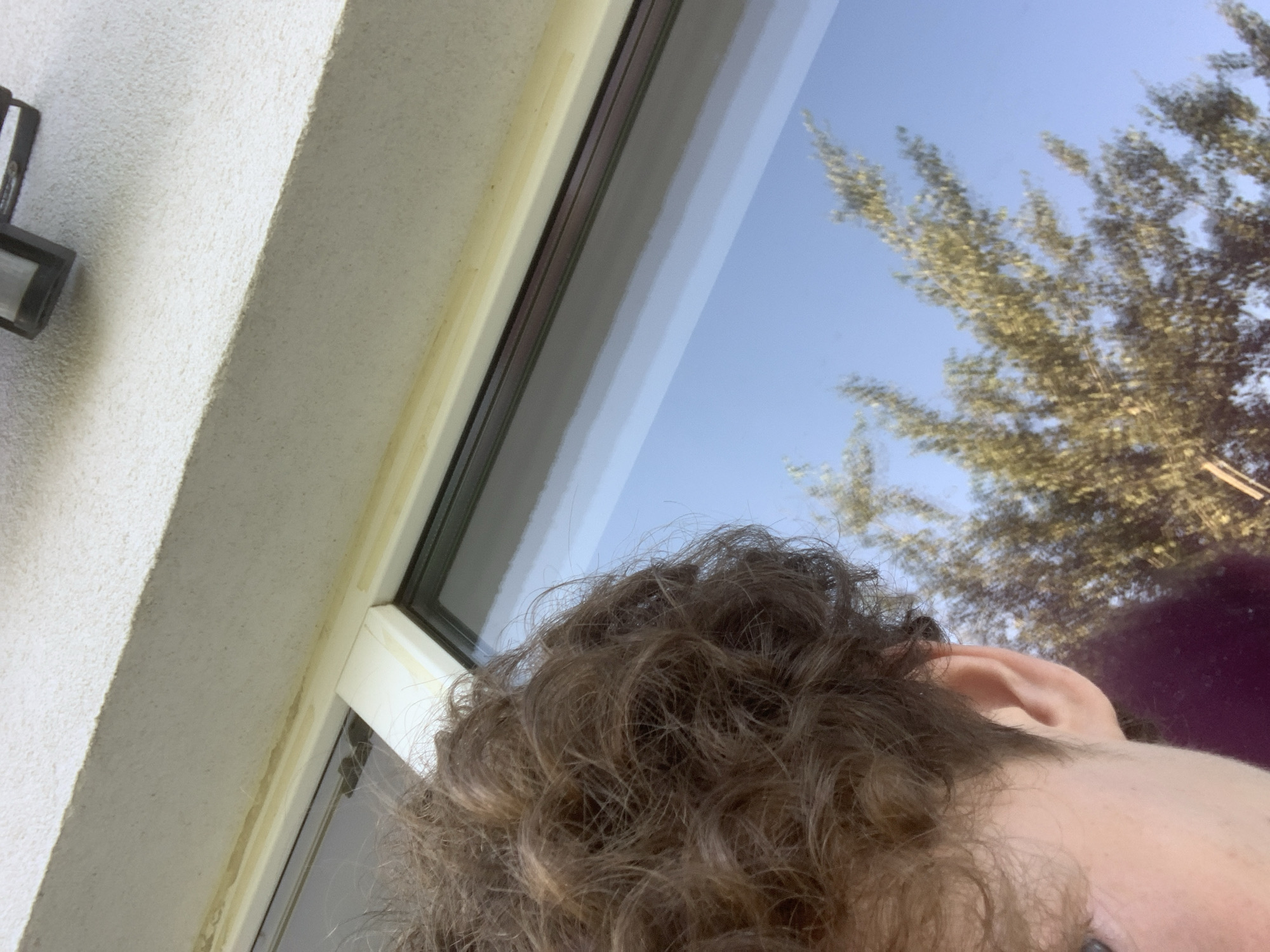 The image is an extreme close-up, low-angle shot of a person's head and ear, with a window and a textured wall in the background. The person has curly brown hair and a pale complexion. The window reflects a clear blue sky and the branches of a tree. A security camera is visible mounted on the wall above the window. The setting appears to be outdoors, possibly on a balcony or near a window on an upper floor of a building. There is no indication of the city of Minsk, Belarus.