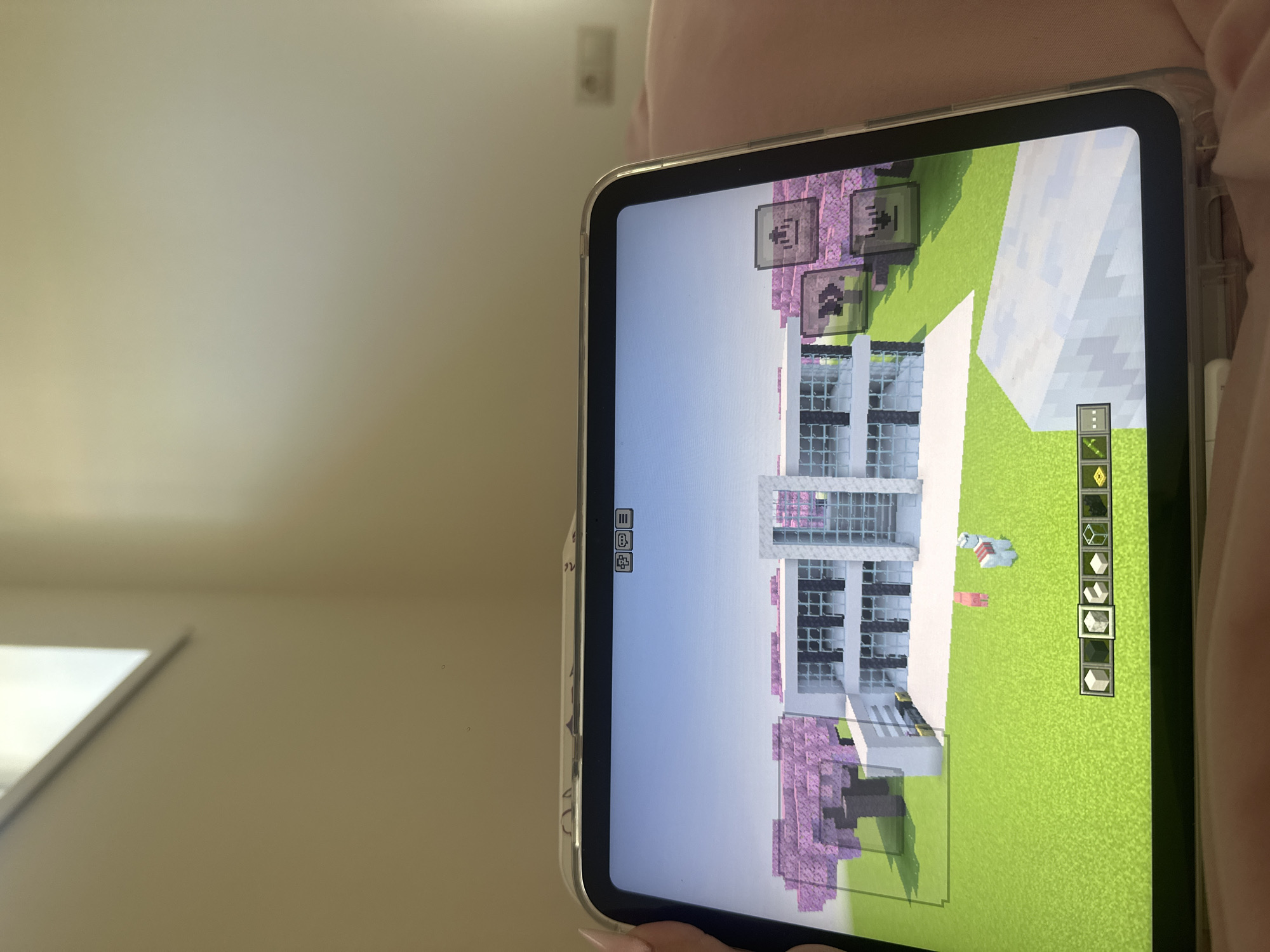 The image shows a tablet displaying a Minecraft game. The game depicts a building with white walls, black-framed windows, and pink trees surrounding it. A green field is visible around the building. The tablet is held in someone's hand. In the background, a wall and a window can be seen. The image does not provide any information to indicate that the location is in Yerevan, Armenia, or any other specific location.