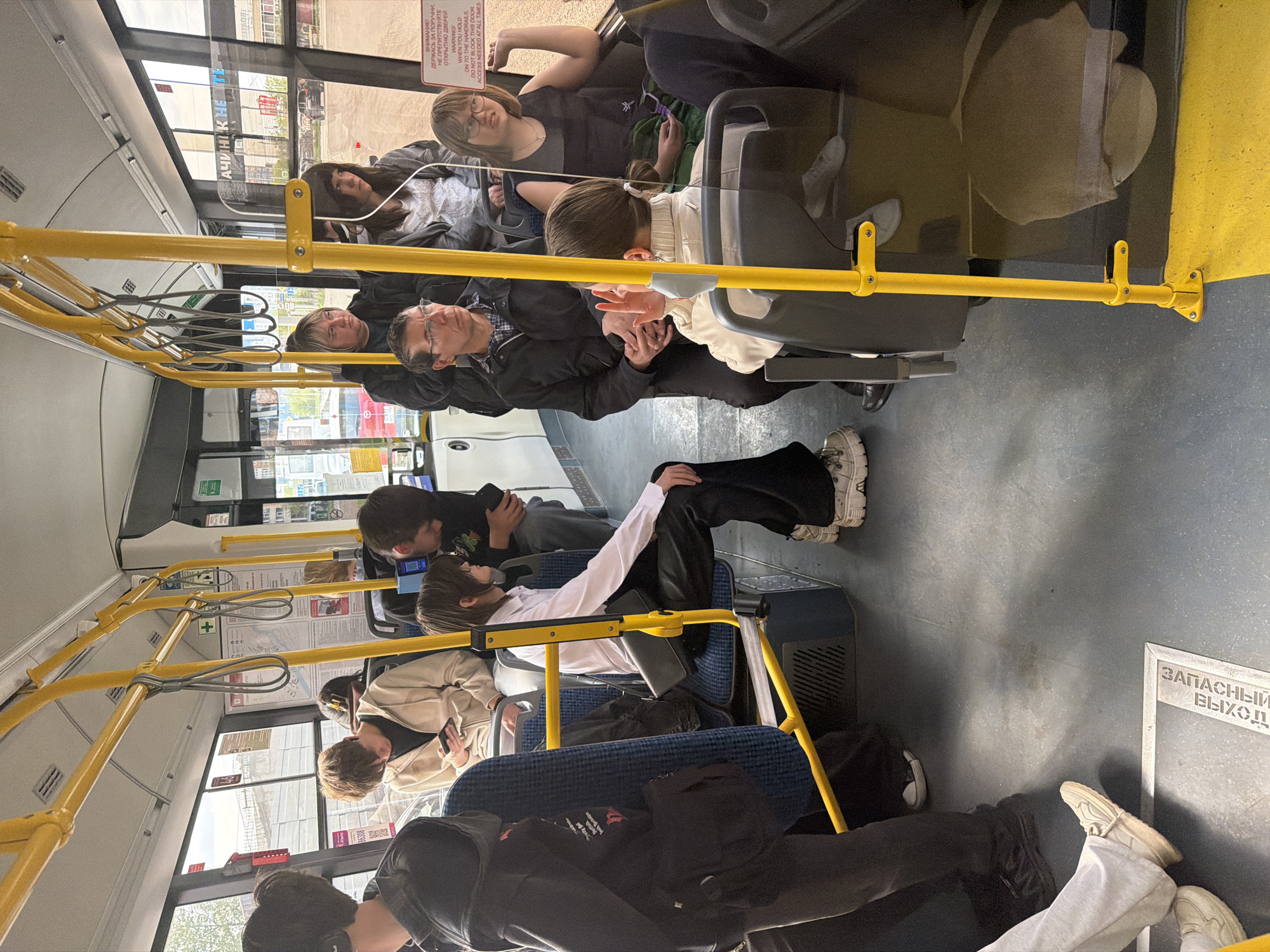 The image captures the interior of a bus in Krasnoyarsk, Russia. Several passengers are seated, most of them looking at their phones. The scene includes the yellow handrails typical of public transportation and the emergency exit sign "ЗАПАСНЫ ВЫХОД". A warning sign in Russian and English instructs people to hold on to the handrails and not block the doors.