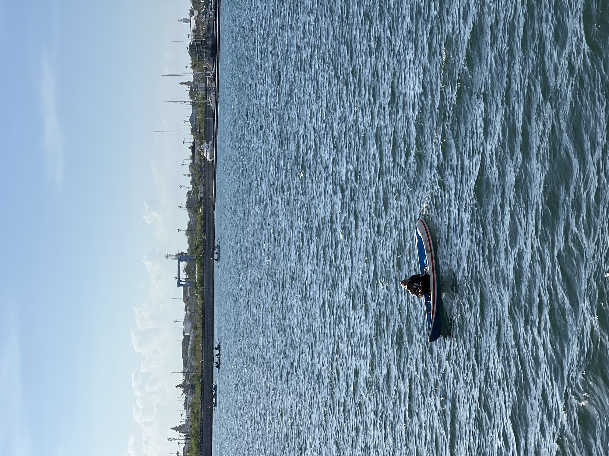 A person wearing a hooded jacket sits in a small blue and white boat on the ocean. The boat has a red stripe along its side. The water is choppy, with small waves. In the background, the coastline of Kuta, Indonesia is visible, featuring a seawall, palm trees, buildings, and several other small boats on the water. Further in the distance, a harbor with several yachts and cranes can be seen. The sky is clear with scattered white clouds.