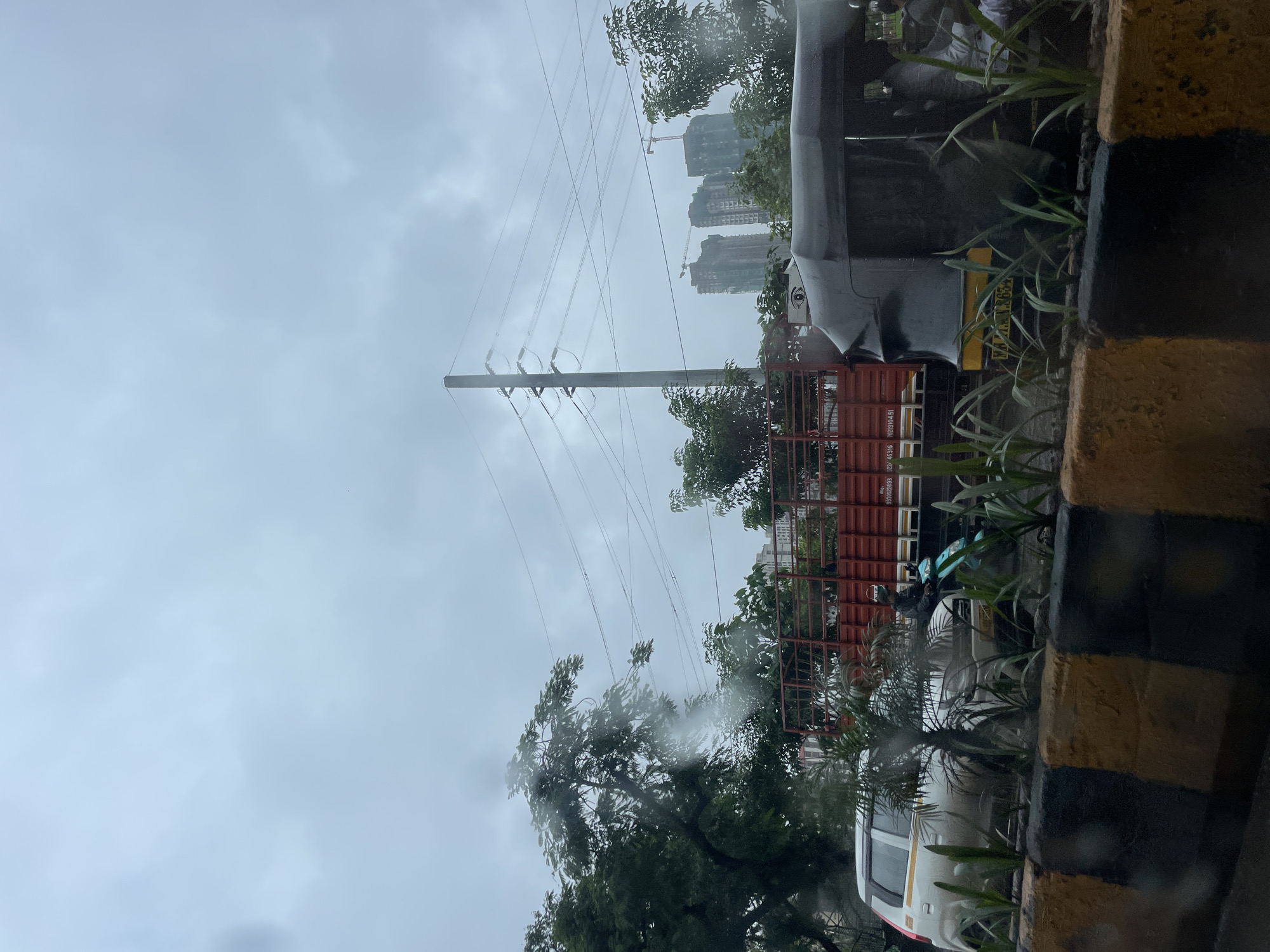 This is a medium shot of a street scene in Mumbai, India, on a cloudy and possibly rainy day. The foreground is dominated by a low concrete barrier with alternating yellow and black painted sections. Behind the barrier, a white car is visible on the left, and a black auto-rickshaw is on the right. A red truck with a metal frame is positioned behind the car and auto-rickshaw. A person is riding a light blue scooter, partially obscured by the car. Green plants line the barrier. In the background, several tall buildings, some appearing to be under construction, are visible. A utility pole with power lines stretches across the frame. The sky is overcast with a grayish-blue hue.