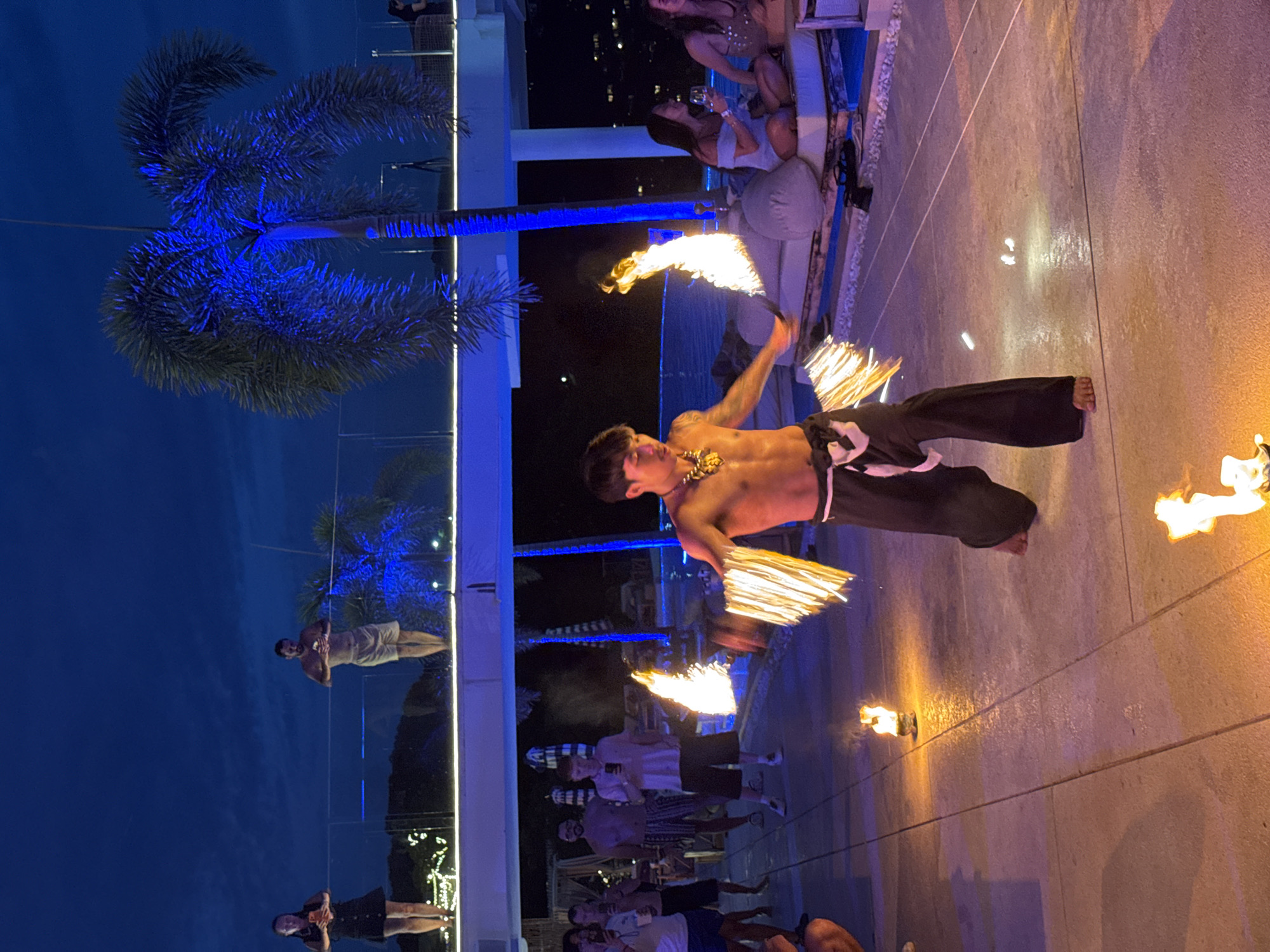 In Karon, Thailand, at night, a male fire dancer performs in front of an audience. The dancer, bare-chested and wearing dark pants, spins flaming props in his hands, creating arcs of light. The ground is paved with large stone tiles, and several small fires burn on the ground around the dancer. In the background, people are visible, some standing and watching, others seated on lounge furniture near a pool. Palm trees are lit with blue light, and the sky is dark blue. The overall scene suggests an outdoor evening event or performance at a resort or beach club.