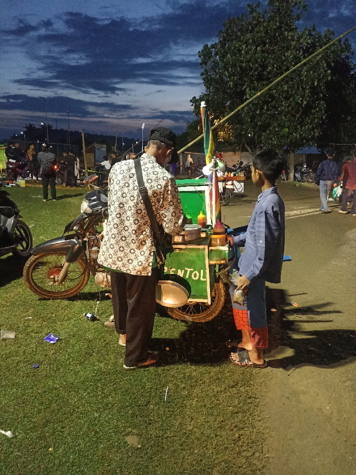 This outdoor image, taken in Kecamatan Pujut, Indonesia, depicts an evening scene with a food vendor and a young customer. The primary subjects are an adult man, seen from behind, and a young boy, facing the vendor. The man wears a patterned long-sleeved shirt (possibly batik), dark pants, and a dark cap, with a bag slung over his shoulder. He is standing behind a green food cart, which appears to be attached to a modified motorcycle. The boy, dressed in a blue long-sleeved shirt and red and blue shorts, stands barefoot or in sandals, holding a plastic bag, looking at the vendor.
The food cart has several bottles of sauces (ketchup, chili sauce) on its counter and is adorned with colorful ribbons or flags. The word "PENTOL" is visible in white letters on the side of the cart, indicating the type of food being sold, which are Indonesian meatball snacks. The motorcycle part of the cart is parked on a grassy area, where some litter (plastic wrappers, a straw) is scattered. The boy stands on a dirt path next to the grass.
In the background, under a twilight sky with dark clouds, numerous other people are gathered, suggesting a community event, market, or fair. Several motorcycles are also visible, parked on the grass or nearby. Large trees frame the scene, and artificial lighting illuminates the immediate area around the food cart, creating long shadows. The overall atmosphere suggests a casual evening gathering.