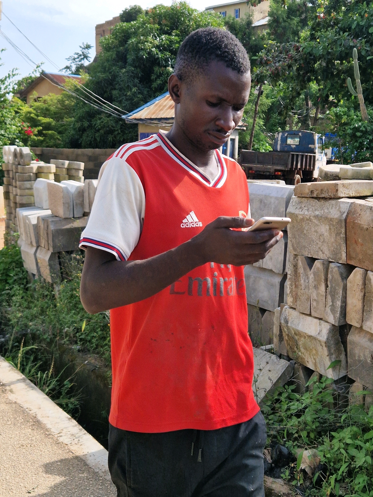 A young Black male wearing a red and white Adidas t-shirt and dark pants is standing outdoors, looking at his smartphone. He is positioned to the right of the frame, with his body angled slightly away from the camera. The background shows a variety of elements, including stacks of concrete blocks, lush green vegetation, a blue truck, and buildings in the distance. The scene appears to be in an urban or semi-urban environment, possibly a construction site or a residential area with building materials stored nearby. The lighting suggests it is daytime, with natural sunlight illuminating the scene.