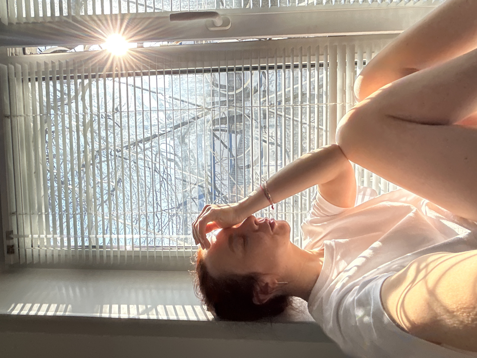 A person lies on a window sill with legs raised, facing towards the window. The window has white blinds, behind which bare trees are visible. Sunlight streams through the window, creating bright highlights and strong shadows on the person and the window sill. The person is wearing a light-colored T-shirt. The location is likely a residence in Chaikovsky, Russia, given the prompt.