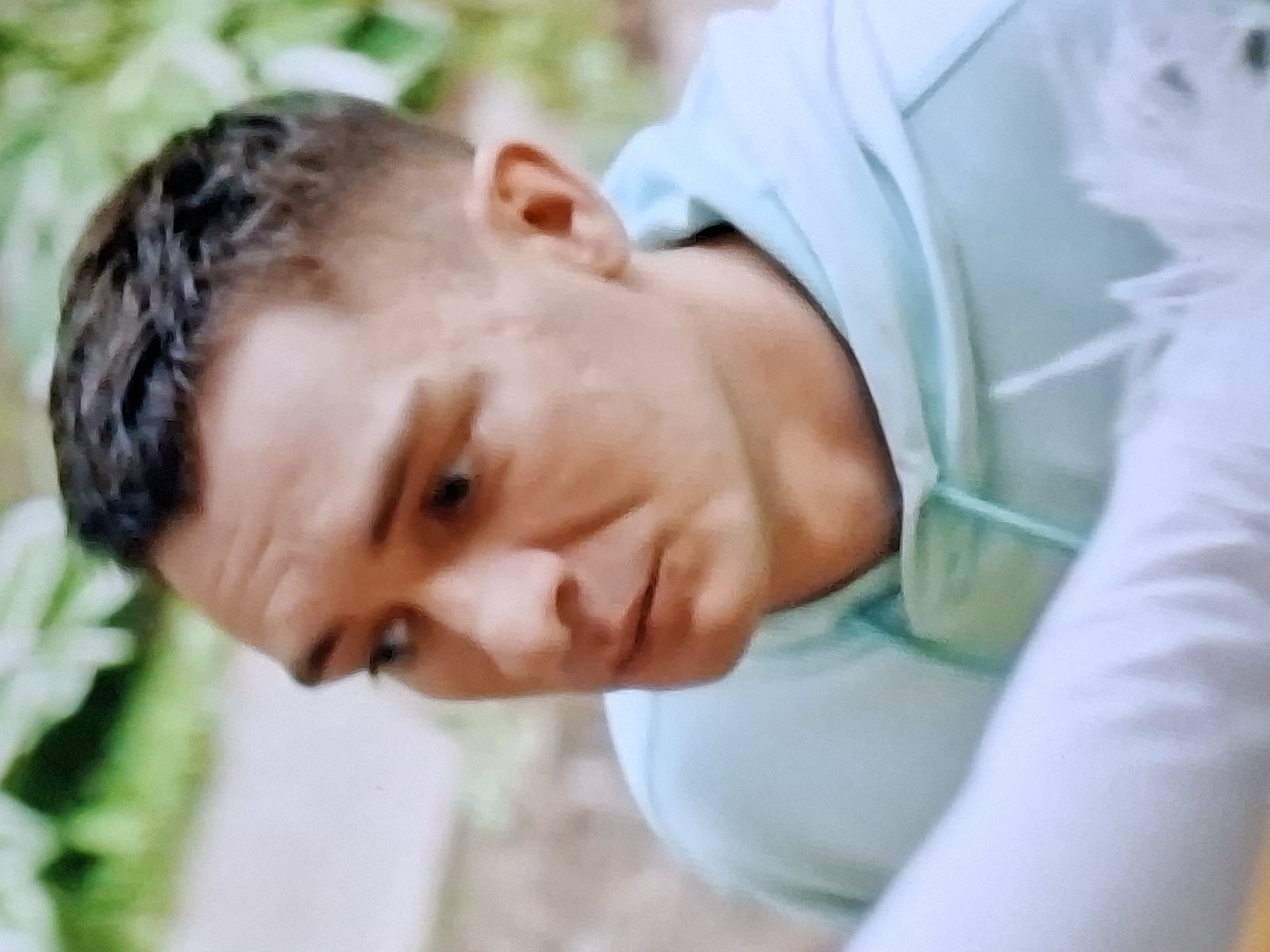 The image is a close-up, portrait-style photograph of a fair-skinned man with short, dark hair. He is wearing a light blue hooded sweatshirt. The man's head is turned to his left, and he is looking slightly down and to his left with a serious expression. His face is in profile from the nose to the ear, with his mouth slightly open. The background is blurred and appears to be outdoors, with hints of green foliage and a light-colored surface, possibly a wall or path. There is no discernible context related to the city of Ivanovo, Russia.