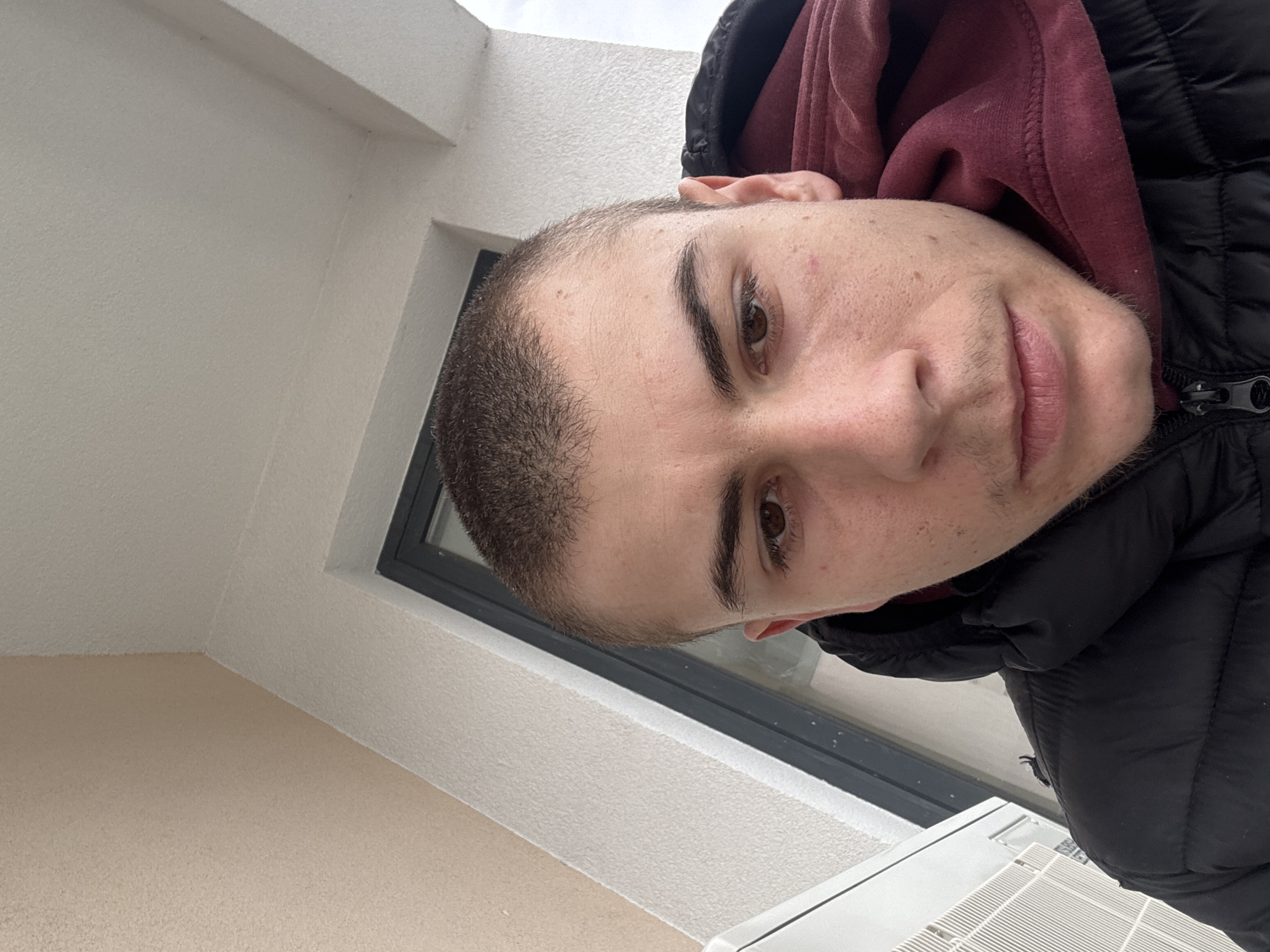 A close-up, angled portrait of a young man with short, dark hair, light skin, and brown eyes. He is wearing a dark-colored puffer jacket with a maroon hoodie underneath. The background suggests an outdoor location, potentially a balcony or patio, with a window or doorway visible behind him. The wall surfaces appear to be textured and painted in neutral tones. The presence of what appears to be an air conditioning unit towards the bottom of the frame hints at an urban setting. It's not possible to confirm the location in Plovdiv, Bulgaria, without further information or recognizable landmarks.