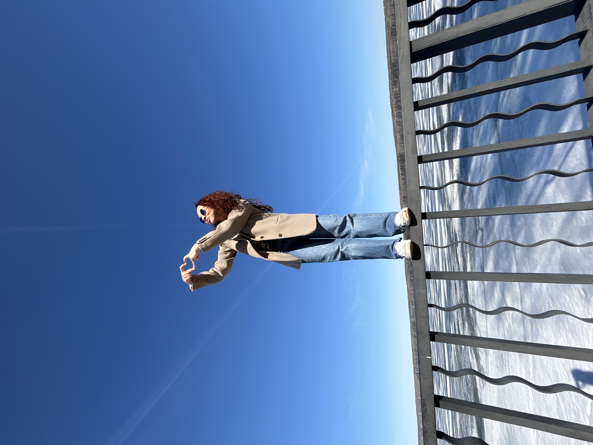A vibrant outdoor, full-length shot captures a person standing next to a body of water under a clear blue sky in Chaikovsky, Russia. The individual, appearing to be female, has curly reddish-brown hair and is wearing dark sunglasses, a light brown or tan blazer, blue jeans, and white sneakers. They are positioned slightly to the left side of the frame, facing towards the right, with both hands brought together to form a heart shape in front of their chest, conveying a gesture of affection or joy.
To the right of the person, a dark metal railing with distinctive wavy horizontal bars stretches across the scene, providing a boundary to a large expanse of water. The water surface, reflecting the bright sky, shows ripples and light reflections, suggesting movement or currents. Above the railing and the person, a vast and clear blue sky dominates the background, exhibiting a subtle gradient from a deeper blue at the top to a lighter shade closer to the horizon. A faint, thin white contrail is visible diagonally across the upper left portion of the sky. The bright illumination indicates a sunny day with excellent weather conditions. There is no visible text in the image.