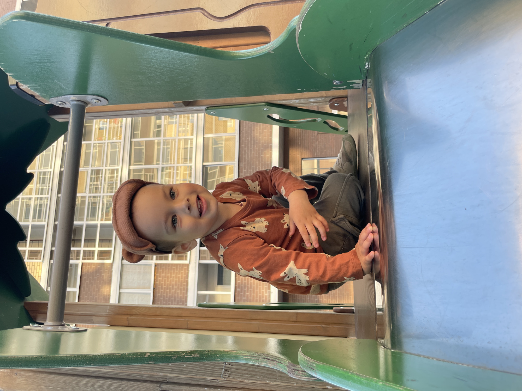 A young child is positioned at the top of a playground slide, located in the city of Izhevsk, Russia. The child, with light brown hair and blue eyes, wears a brown fabric hat with two small ear-like protrusions, an orange long-sleeved shirt patterned with white animals, dark olive-green pants, and light-colored sneakers. The child is smiling broadly, facing slightly to the viewer's right, with the left hand resting on the smooth, silver-colored metal surface of the slide. The playground structure surrounding the child consists of green painted wooden or plastic elements, including a curved barrier to the left and a flat panel secured by bolts to a wooden frame on the right, which also features a horizontal metal bar. In the background, a multi-story brick building with numerous white-framed rectangular windows is visible under daylight illumination.