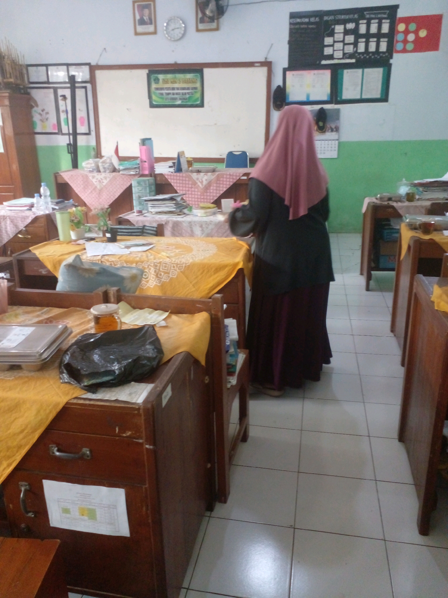The image shows the interior of a classroom in Banyuputih, Indonesia. A person wearing a pink hijab and a dark abaya is standing with their back to the camera, facing a whiteboard. The classroom appears to be furnished with wooden desks and chairs. There are various items on the desks, including books, papers, and bottles of water. On the walls, there are framed pictures, a clock, and a bulletin board with information. The floor is tiled. The overall impression is of an active learning environment.